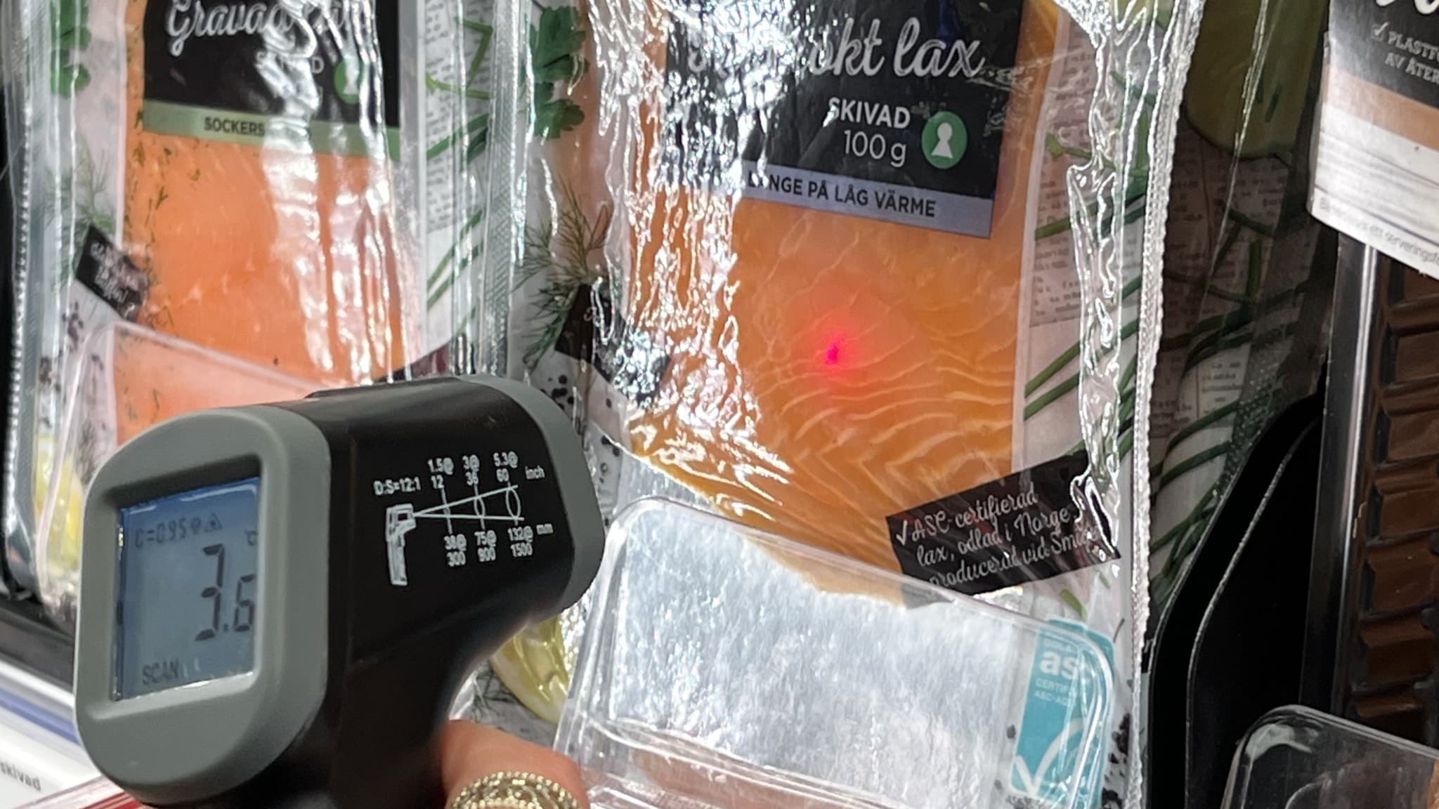 En hand håller en termometer som mäter temperaturen på förpackad lax i en butik. Displayen på termometern visar 3,6 grader Celsius. En röd laserpunkt syns på plastförpackningen.