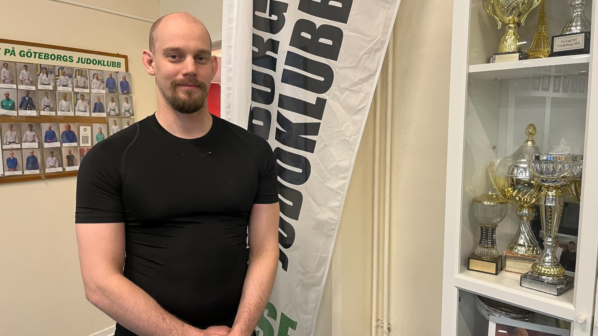 Robin Persson, fallinstruktör, står framför en vit flagga med texten 'Idrottsklubb' samt en vit hylla fylld med pokaler och utmärkelser. En anslagstavla med bilder syns i bakgrunden.