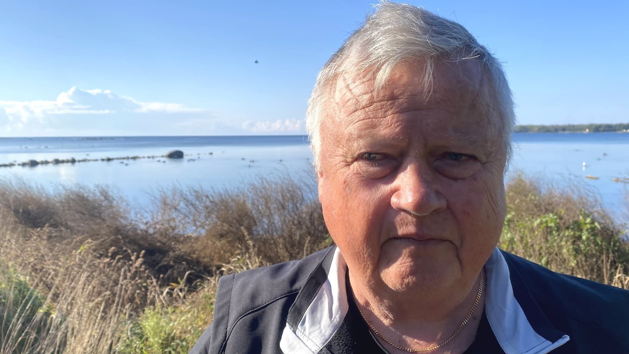 Lennart Arvidsson, fiskare och Mjällbysupporter, står vid en kustlinje med stilla vatten och vass i bakgrunden under en klarblå himmel.