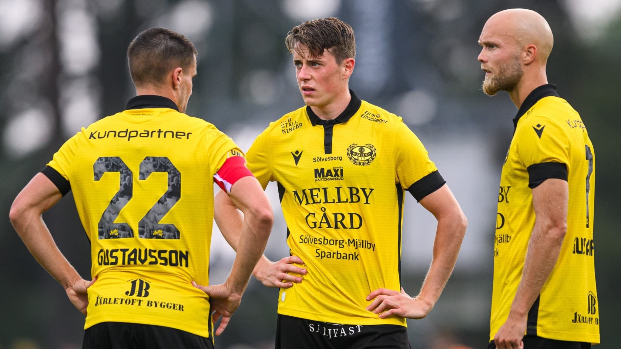 Tre fotbollsspelare i gula tröjor med svart text står på planen och diskuterar under en match. En av spelarna bär nummer 22 och kaptensbindel. Träden syns suddigt i bakgrunden.