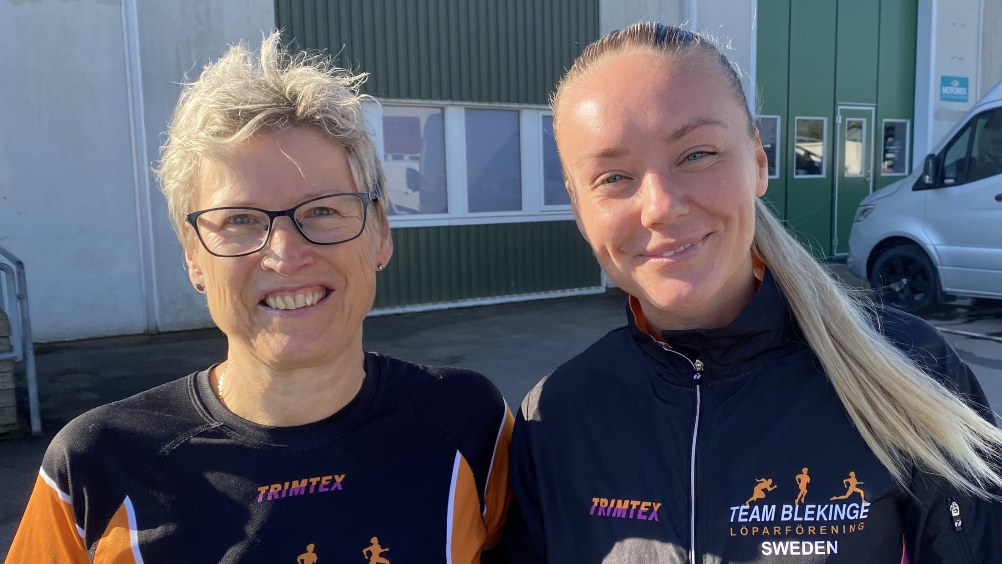 Två personer från Team Blekinge står utomhus framför en industribyggnad, klädda i träningskläder med Team Blekinges logotyp tydligt synlig på tröjorna.