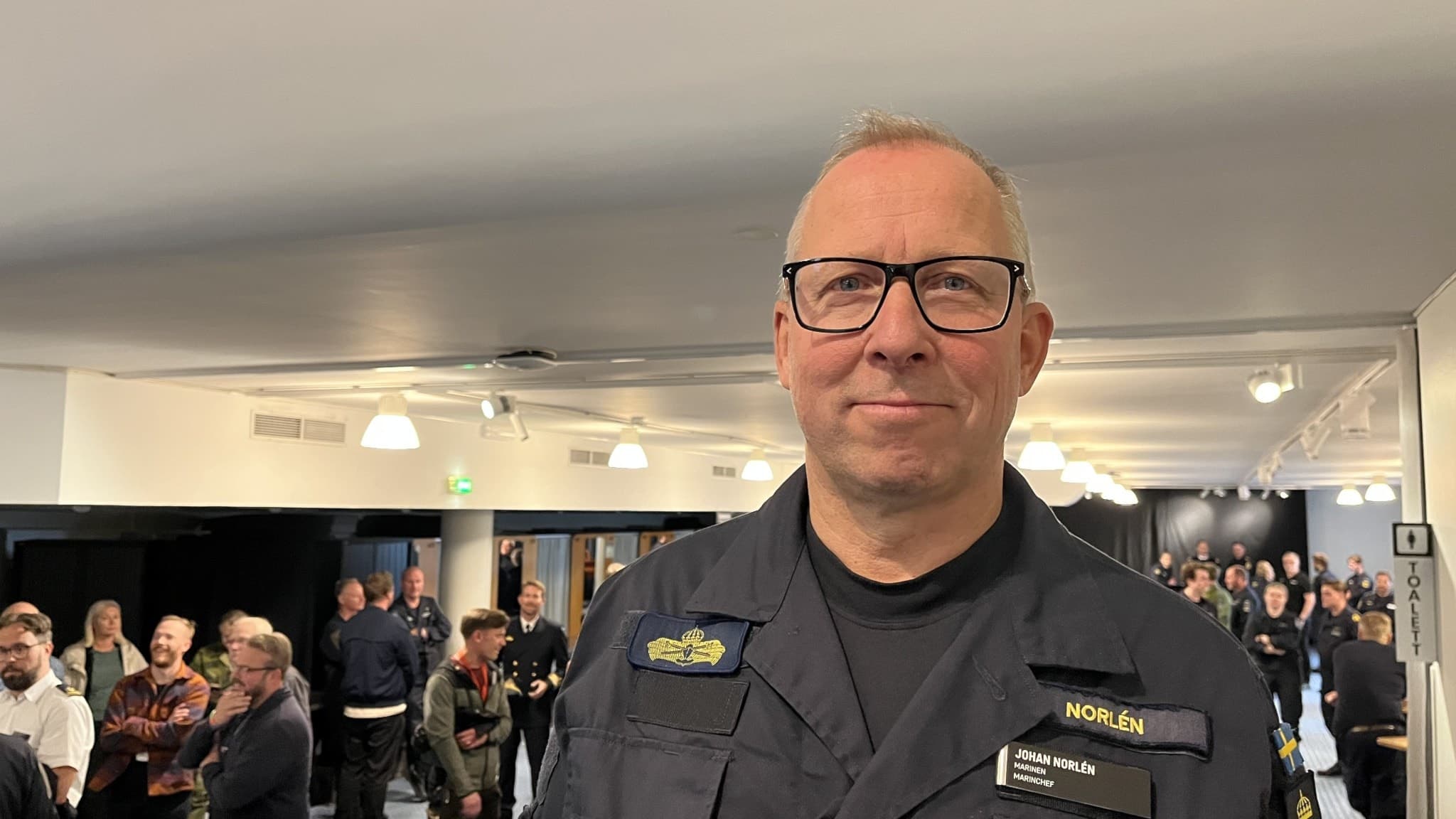 En man i marinuniform står inomhus framför en grupp människor i en samlingslokal. Han bär flera insignier på uniformen och verkar vara i en ledande roll. Gruppen i bakgrunden står i samtal.a
