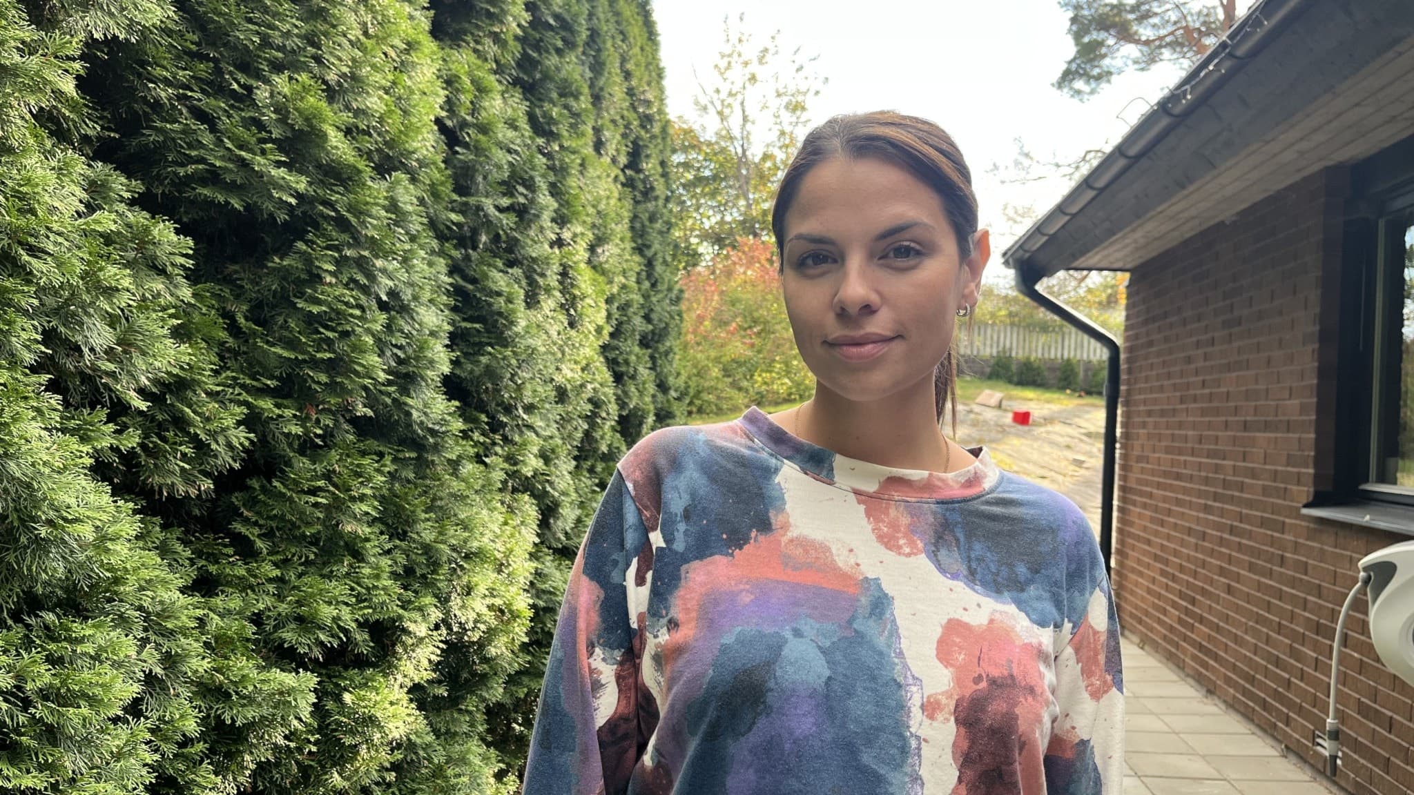 Maria Djozic står utomhus vid en tegelbyggnad, klädd i en färgglad tröja. I bakgrunden syns grönskande buskar och ett staket som delar av området.