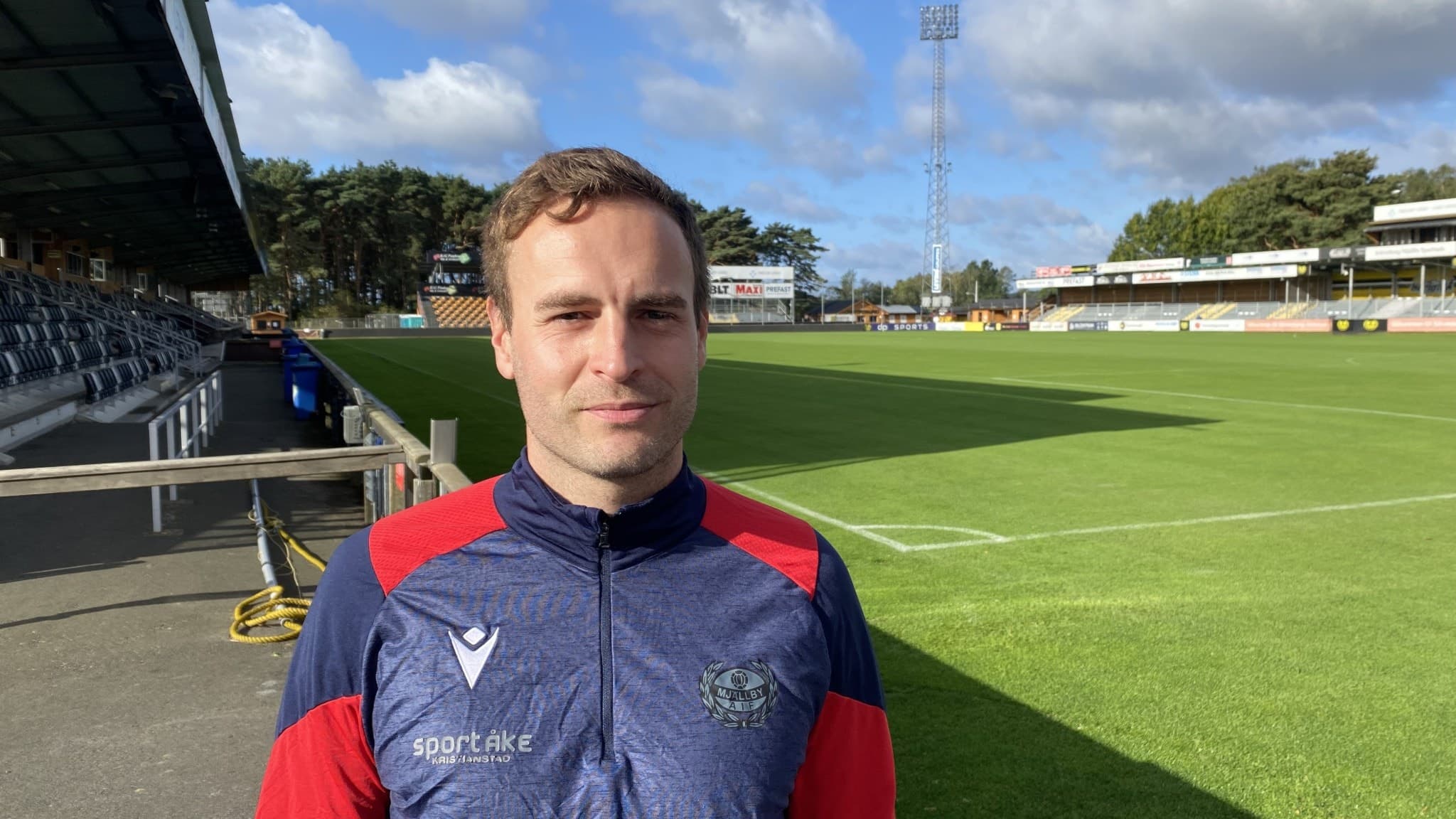 Karl Marius Acksum, tränare för Mjällby, står på en fotbollsarena iklädd en träningsjacka i Mjällbys färger. I bakgrunden syns en grön fotbollsplan, läktare och en klar himmel med vita moln.