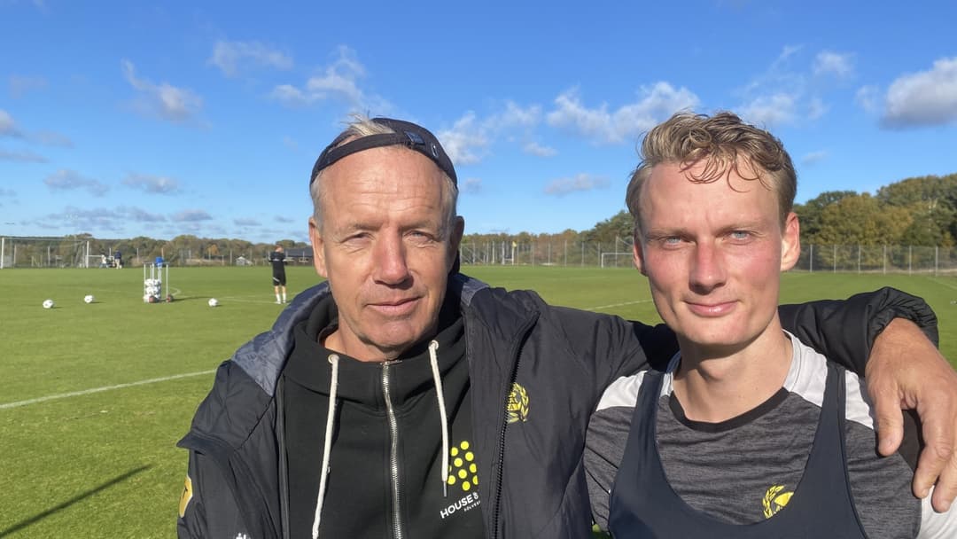 Hasse Larsson och Alexander Johansson står på en grön fotbollsplan under en klarblå himmel. De har på sig träningskläder och ler mot kameran. I bakgrunden syns fotbollsmål och träningsutrustning.
