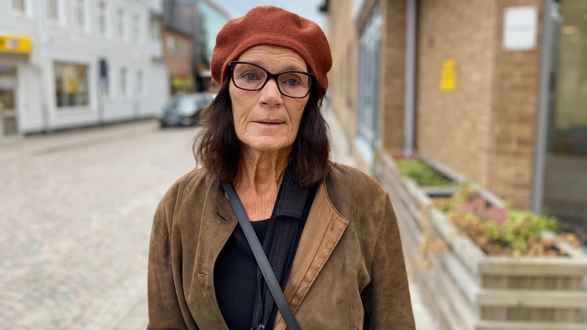 Eva Danielsson står på en stenlagd gata med butiksfasader och byggnader i bakgrunden, klädd i brun kappa och röd basker. Miljön är lugn och typisk för en mindre stad.