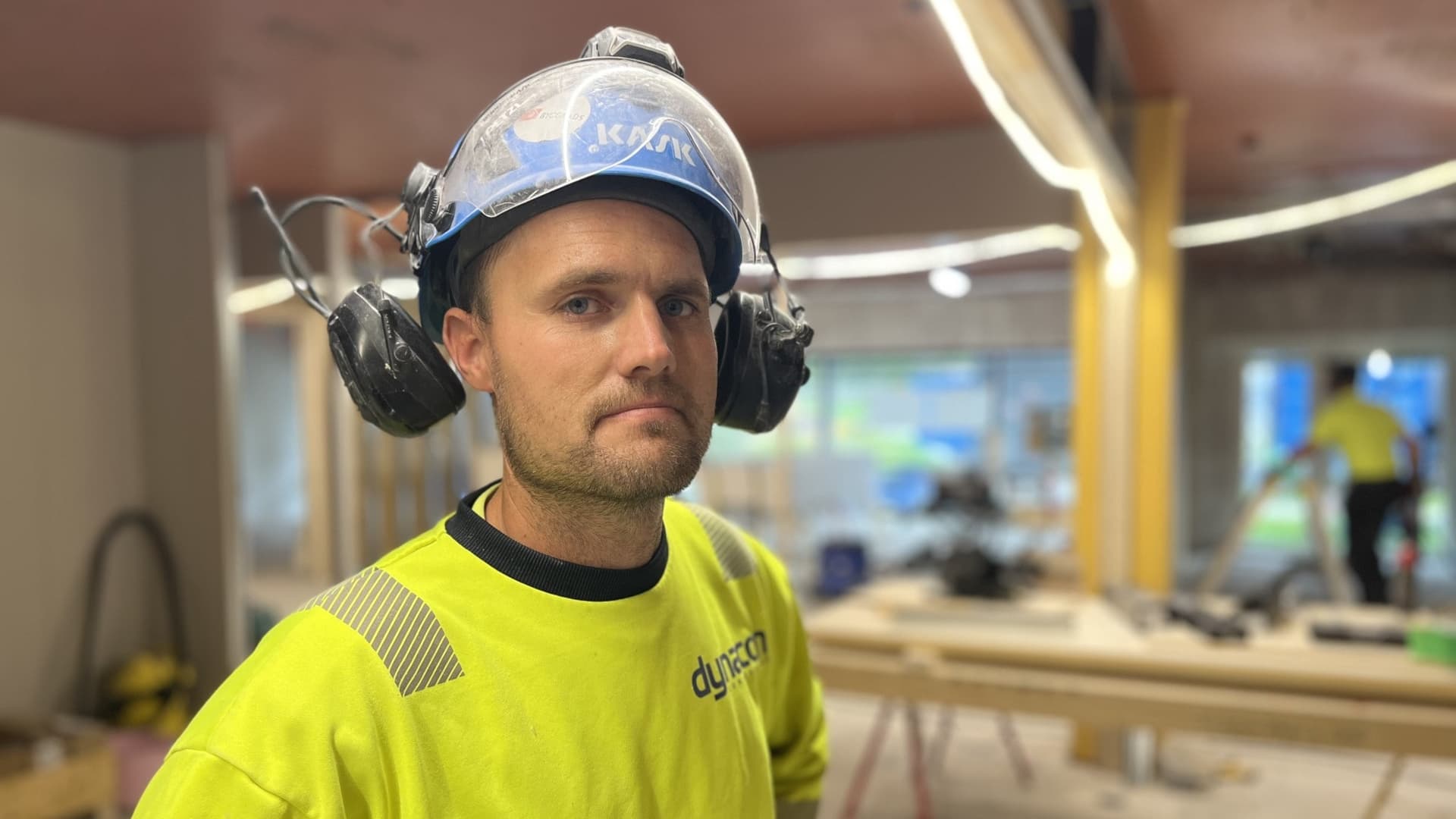 En byggnadsarbetare i gul varseltröja och skyddsutrustning står i ett pågående byggprojekt med träbalkar och verktyg synliga i bakgrunden. Snickaren Michael vill också ha kortare arbetsdag