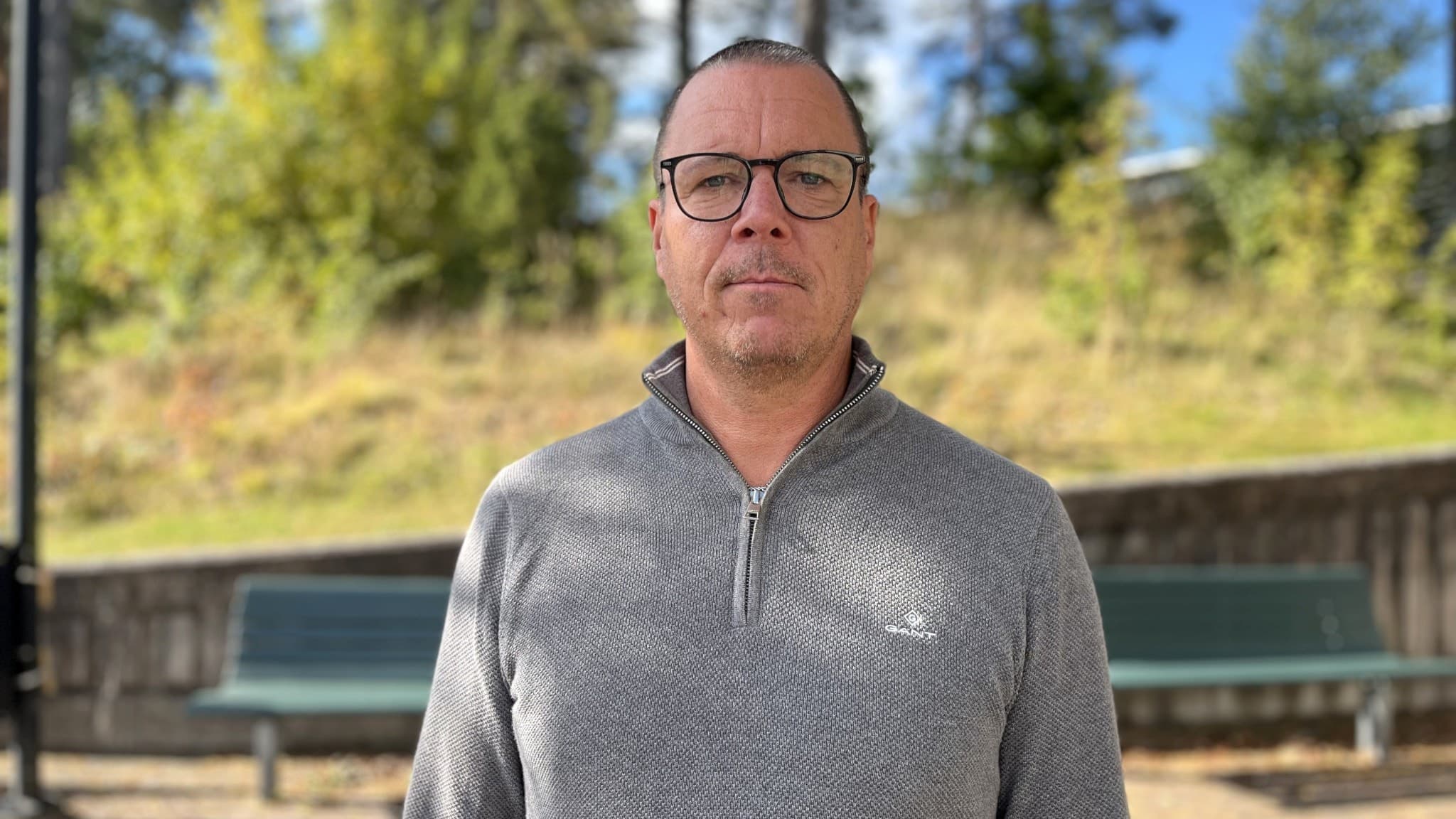 Jimmie Nilsson, skolchef i Ljungby kommun, står utomhus framför en grön bänk med träd och gräskulle i bakgrunden. Han bär en grå stickad tröja och det är soligt väder.
