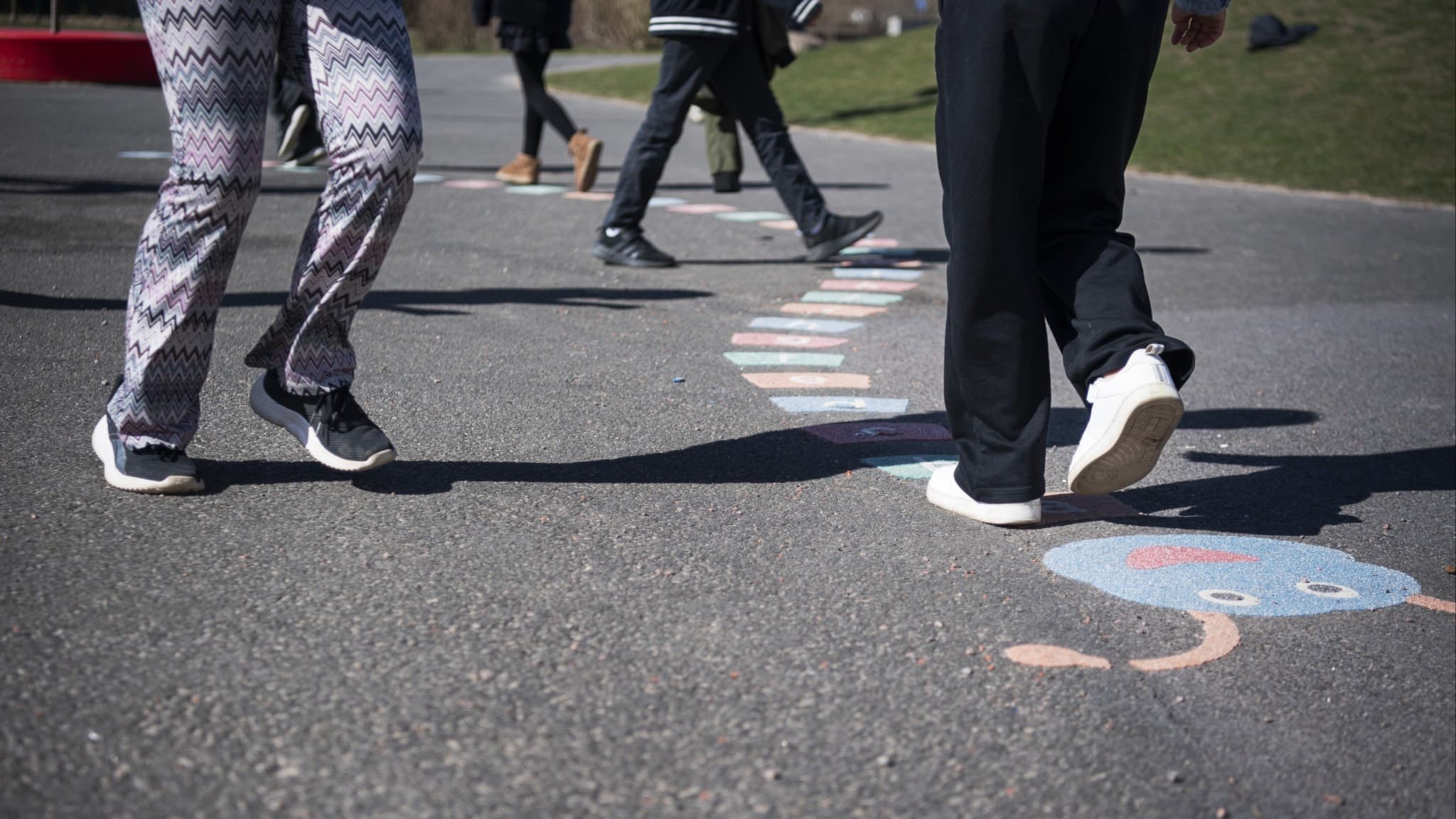 Elever på en skolgård hoppar hage under rasten. Bilden visar benen på barn iklädda färgglada byxor och sneakers. Aktiviteten sker i på en solig dag..