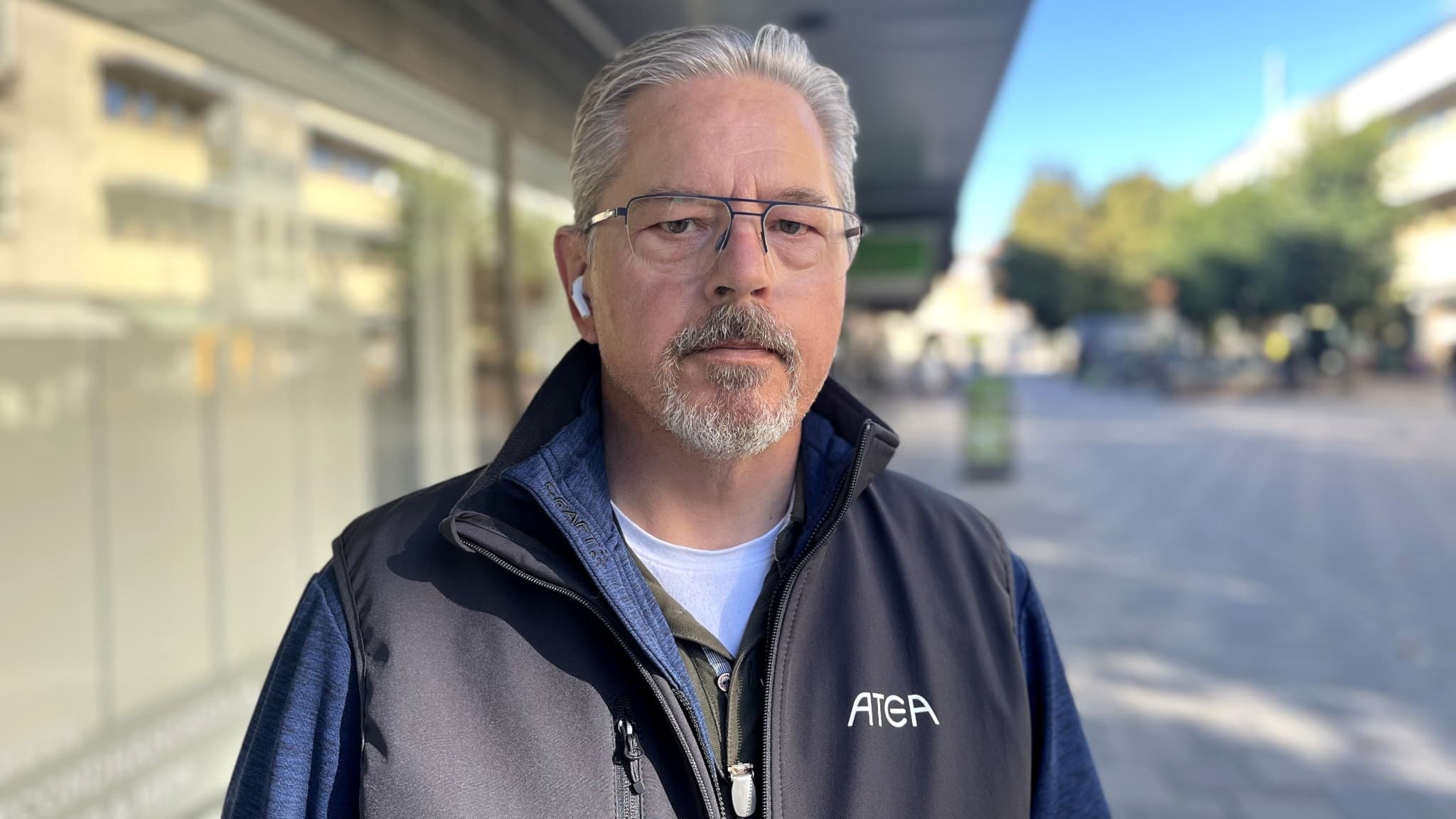En man står utomhus i Växjö framför en modern byggnad och öppet torg. Han har på sig en mörk jacka med texten 'AFR' och kort grått hår syns tydligt. Solen skiner och det är dagsljus.