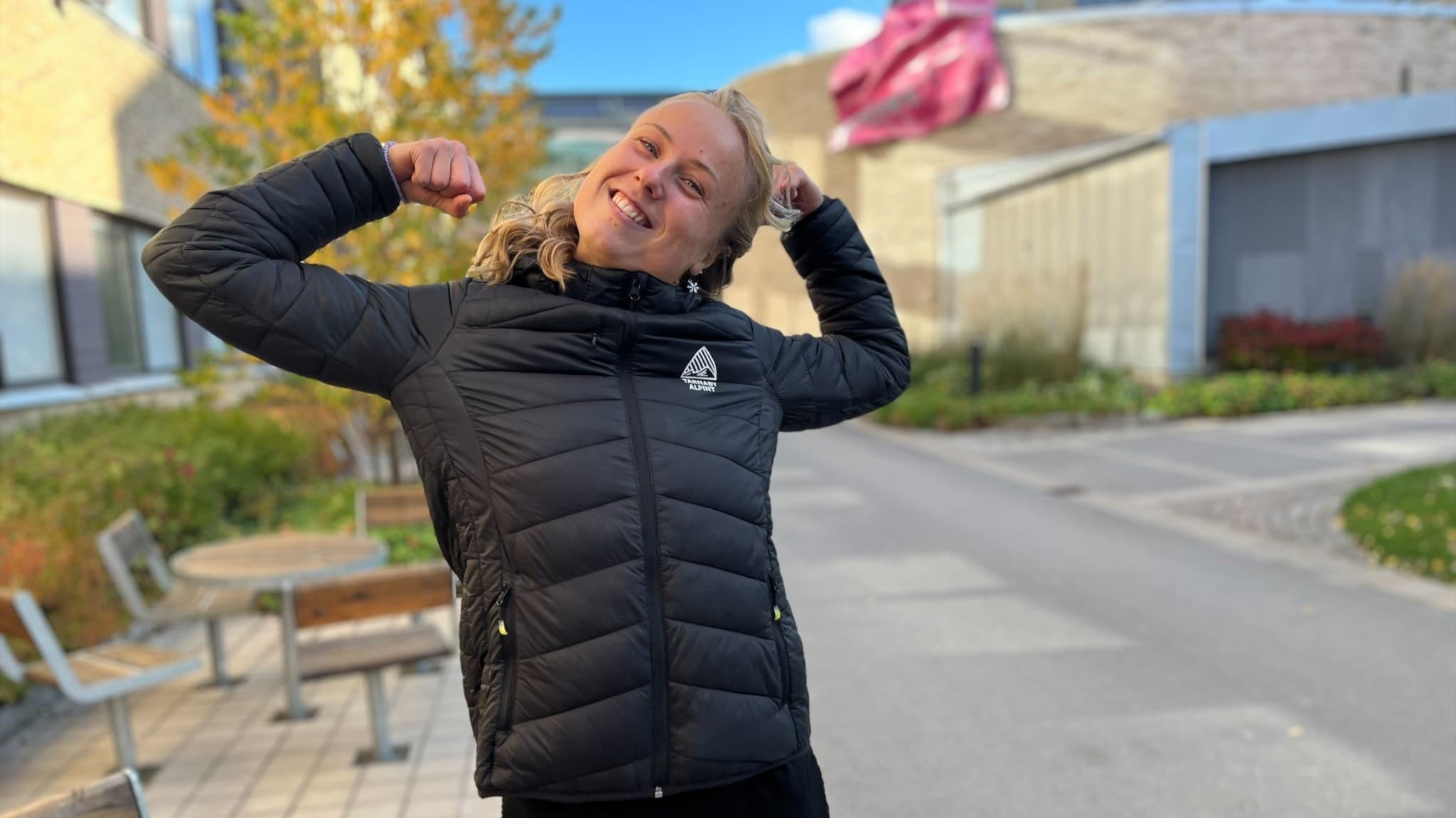 Marceline Forsberg står utomhus i en svart jacka och gör en segergest med båda armarna. Hon förbereder sig inför VM i Ironman på Hawaii. Bakgrunden visar en höstlig miljö med byggnader och träd.