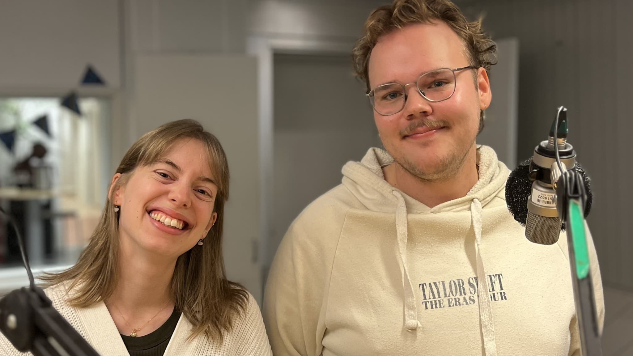 En ung kvinna och en ung man står i en radiostudio med mikrofoner runtomkring. De har ljusa kläder och befinner sig i en professionell inspelningsmiljö.