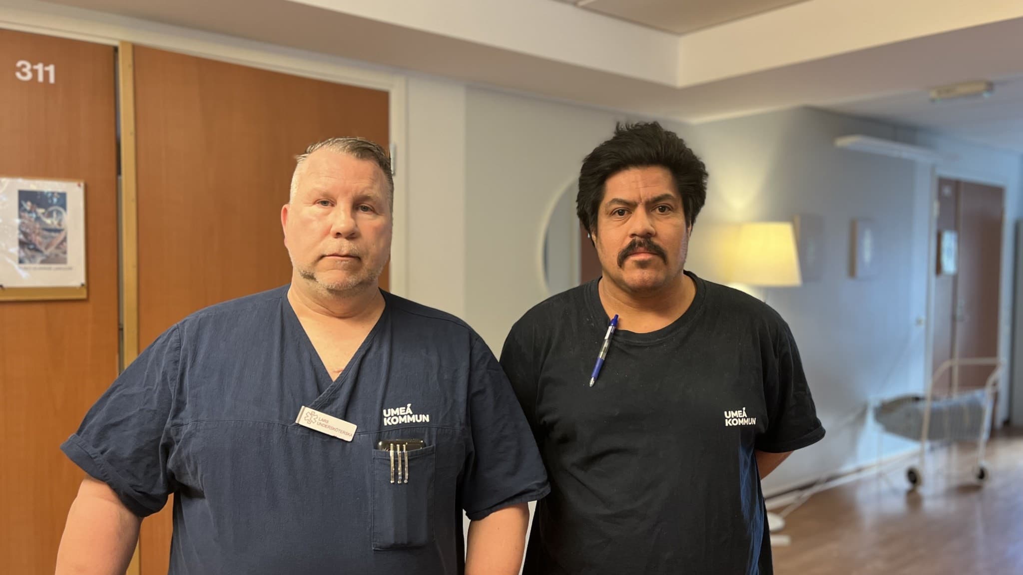 Lars och Sebastian står i en korridor med träfärgade dörrar och ljusa väggar. 