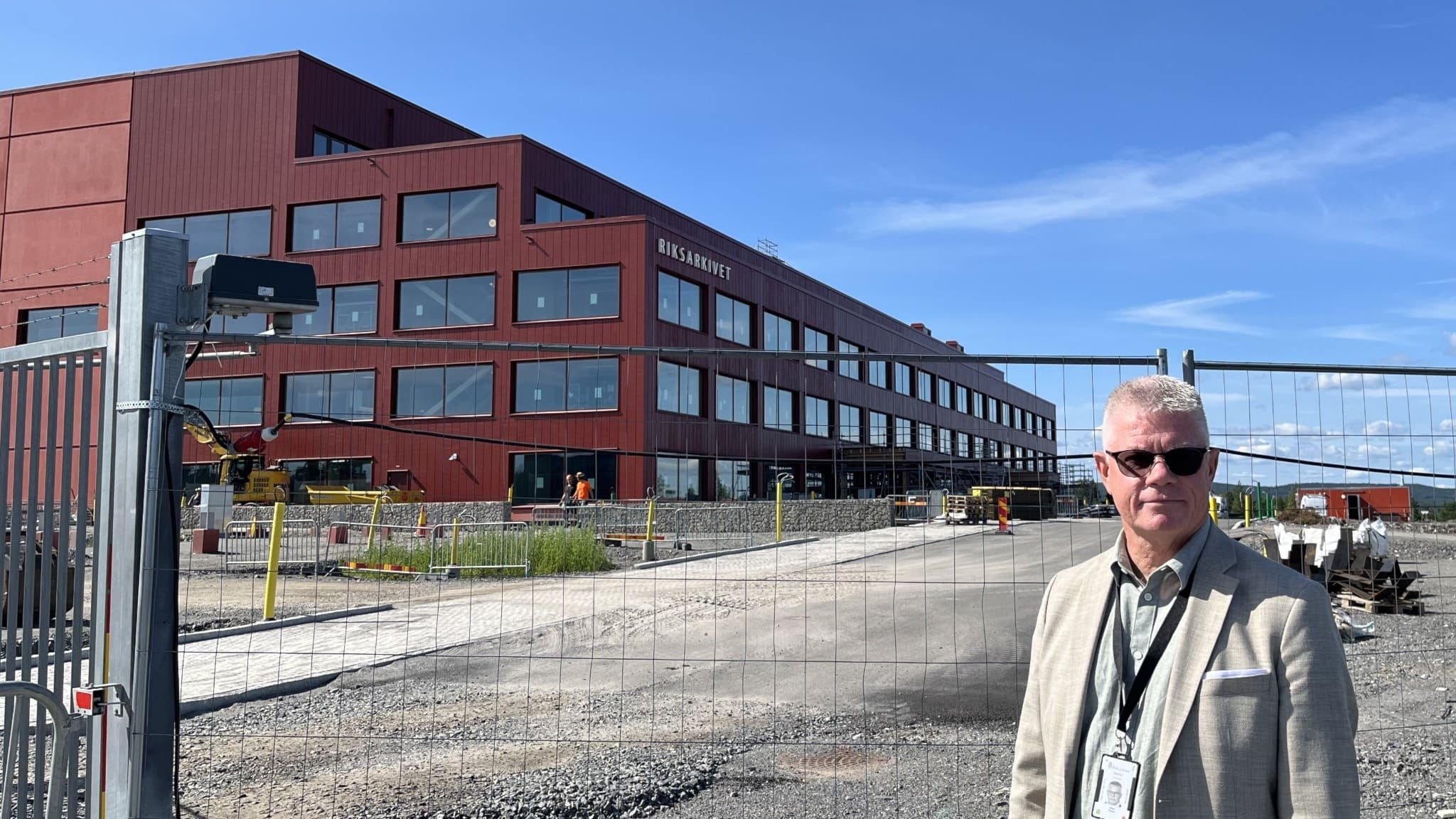 Håkan Viklund står framför en modern röd byggnad med stora fönster, omgiven av ett pågående byggprojekt med grus och arbetsmaterial under en klarblå himmel.