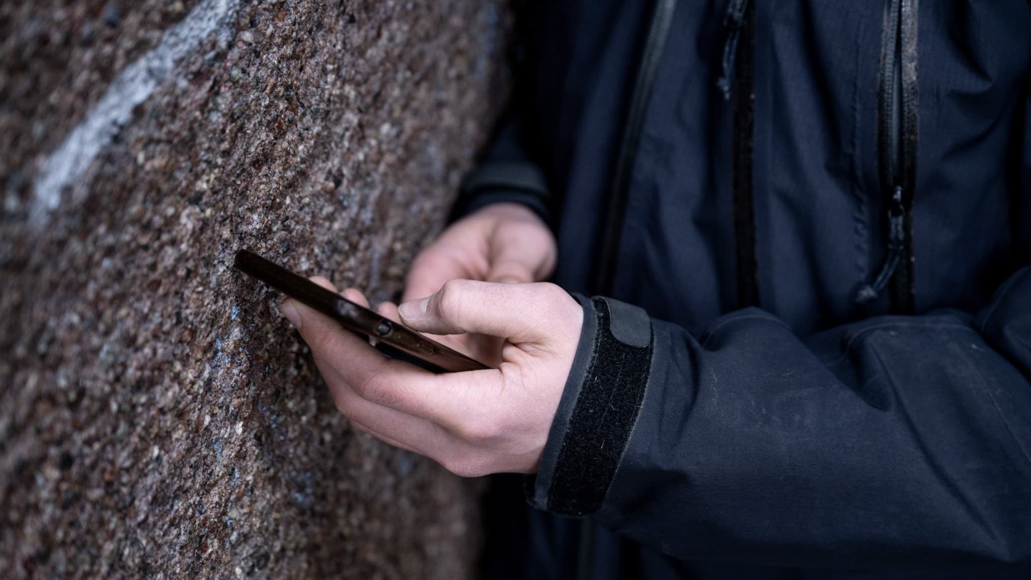 En ung person i mörka kläder står lutad mot en vägg och håller en mobiltelefon i händerna. Fokus ligger på händerna och telefonen, medan ansiktet är utanför bild.