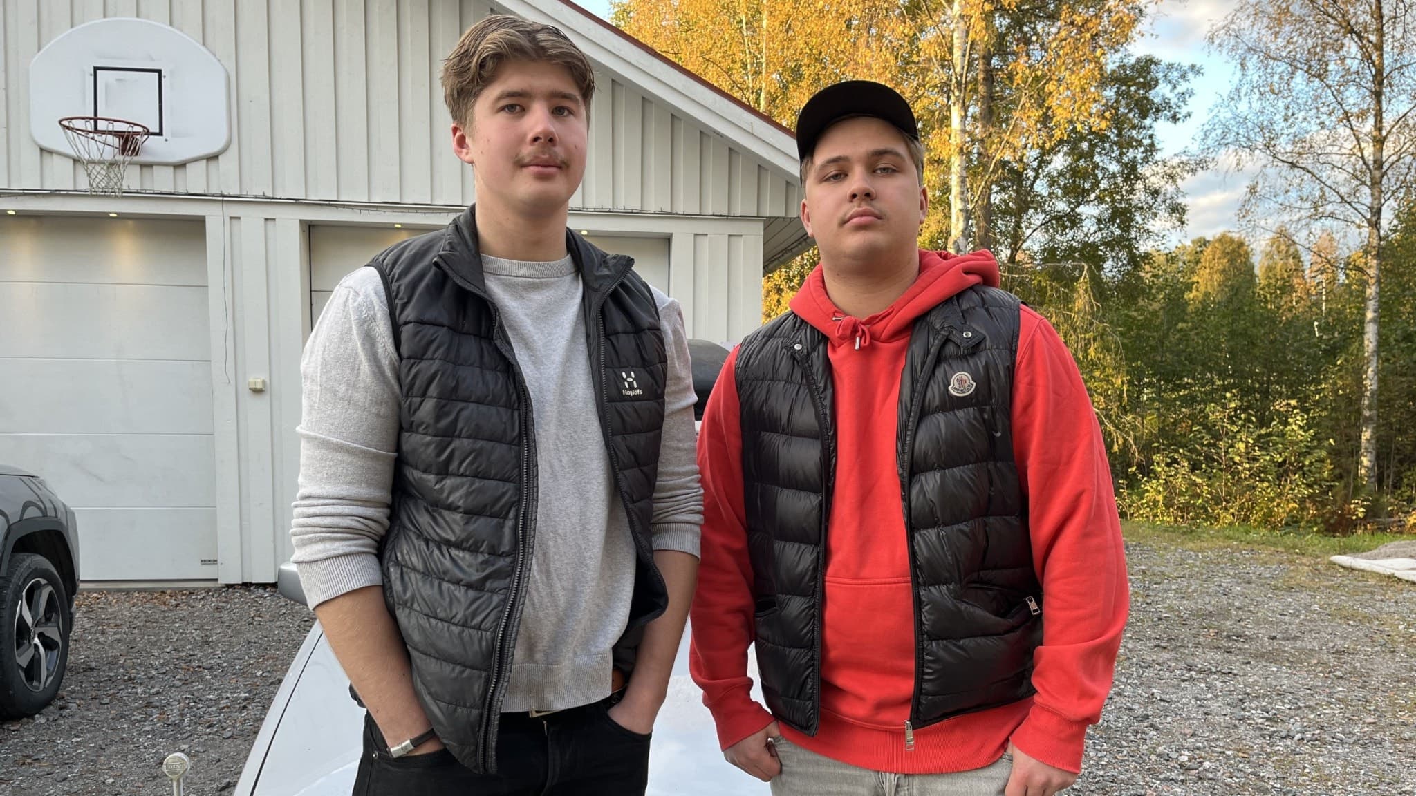 William Olsson Karlsson och Willgot Hirvelä Göransson står utomhus vid en grusplan framför en vit byggnad och en bil, omgivna av höstfärgade träd i Örnsköldsvik.