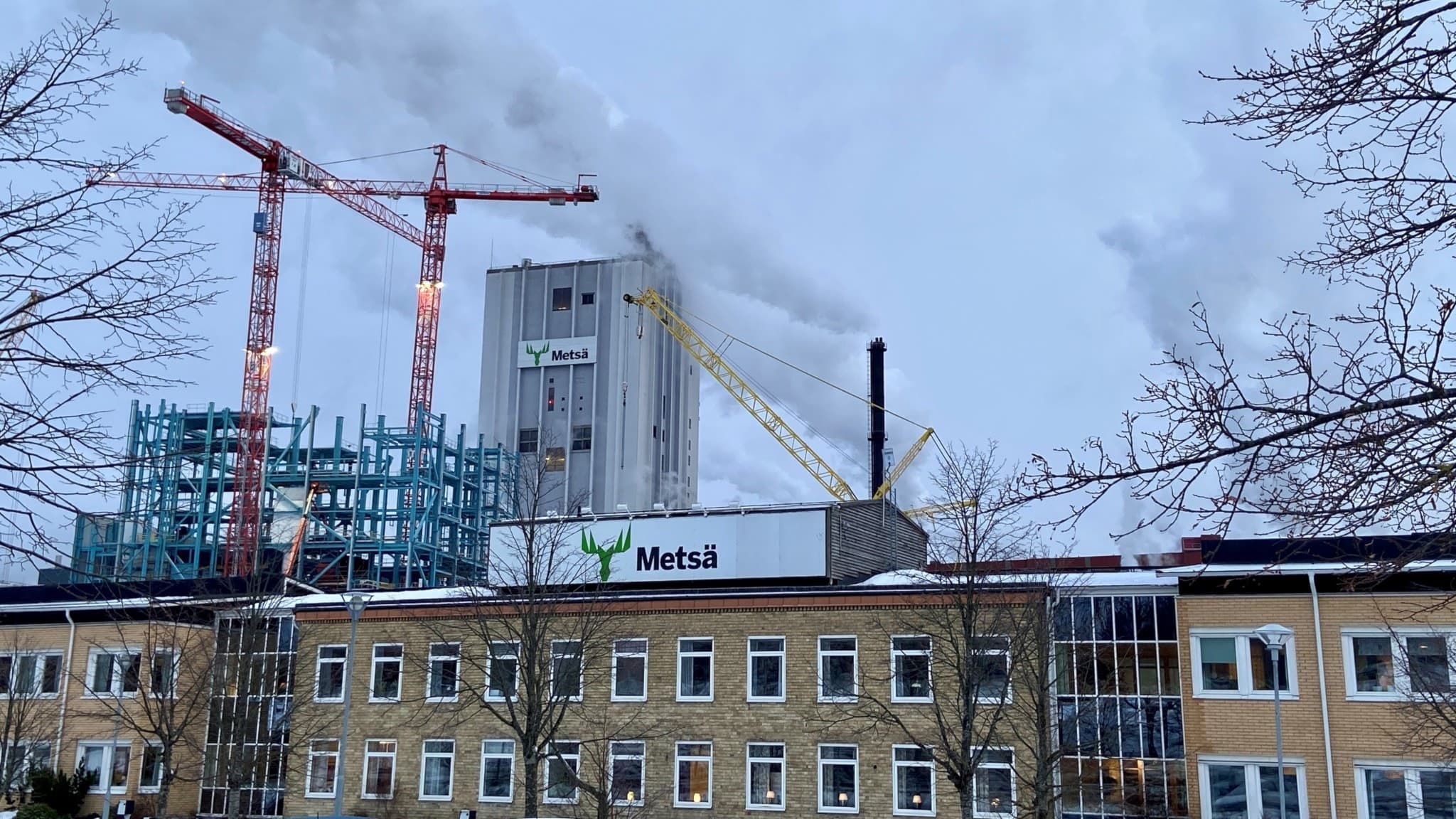 En bild av Husums Fabriker sett framifrån. I förgrunden ser man en lägre gul tegelbyggnad, ovanpå den en skylt med texten "Metsä". I bakgrunden sticker den höga kokaren upp och runt om är det lyftkranar i luften. Foto: Alexander Arvidsson/Sveriges Radio