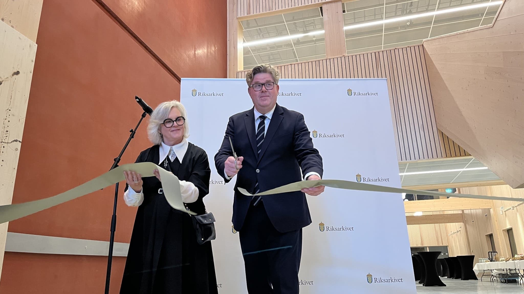Anna Åström Iko, generaldirektör för Riksarkivet, och Gunnar Strömmer (M), justitieminister, klipper gemensamt ett band vid en invigningsceremoni i en modern byggnad med ljus interiör.