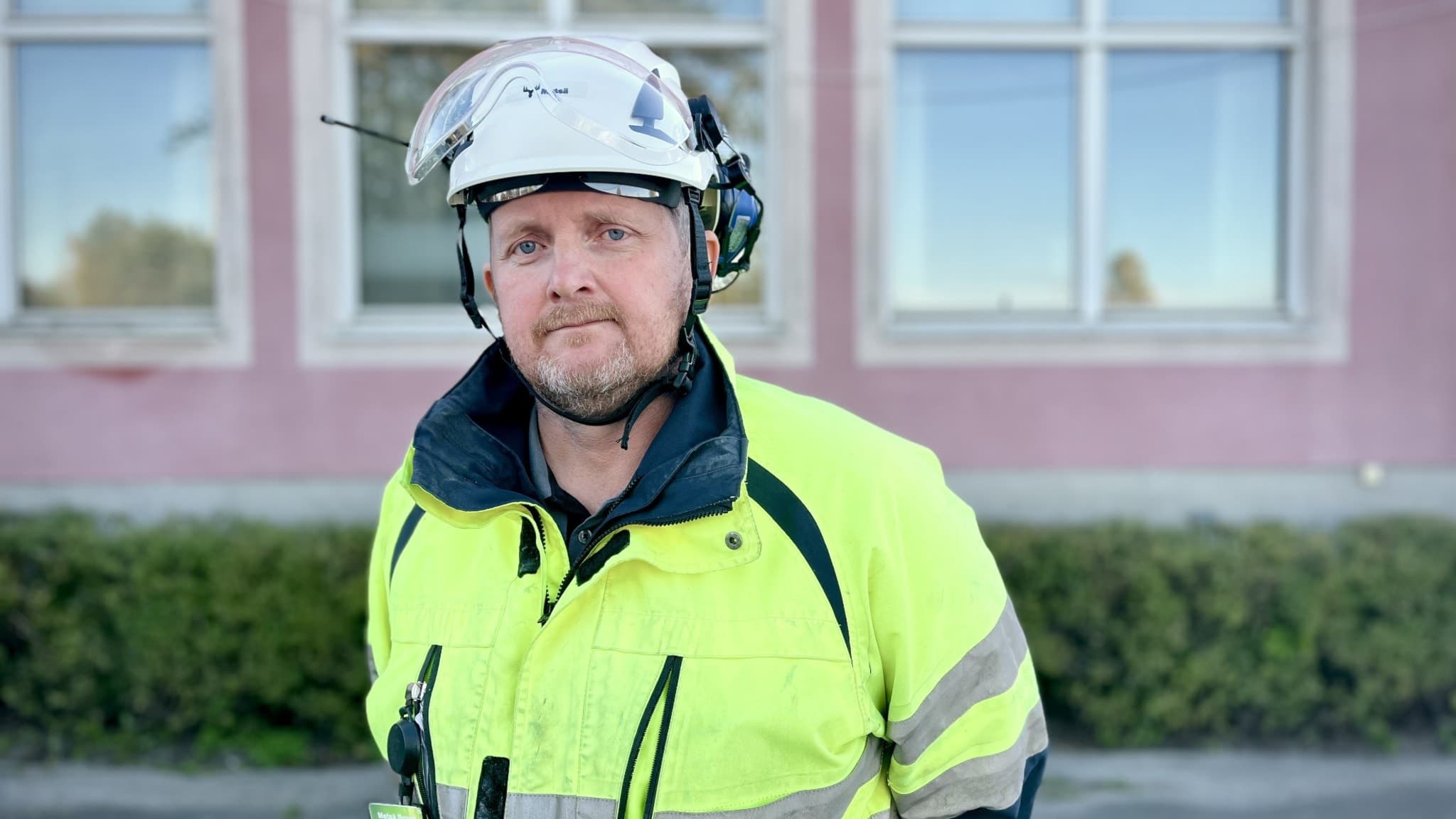 Ulf Häggqvist, ordförande för Pappers Avd 143 Husum, står utomhus i högsynlig arbetsjacka och skyddshjälm framför en byggnad med rosa fasad och stora fönster.