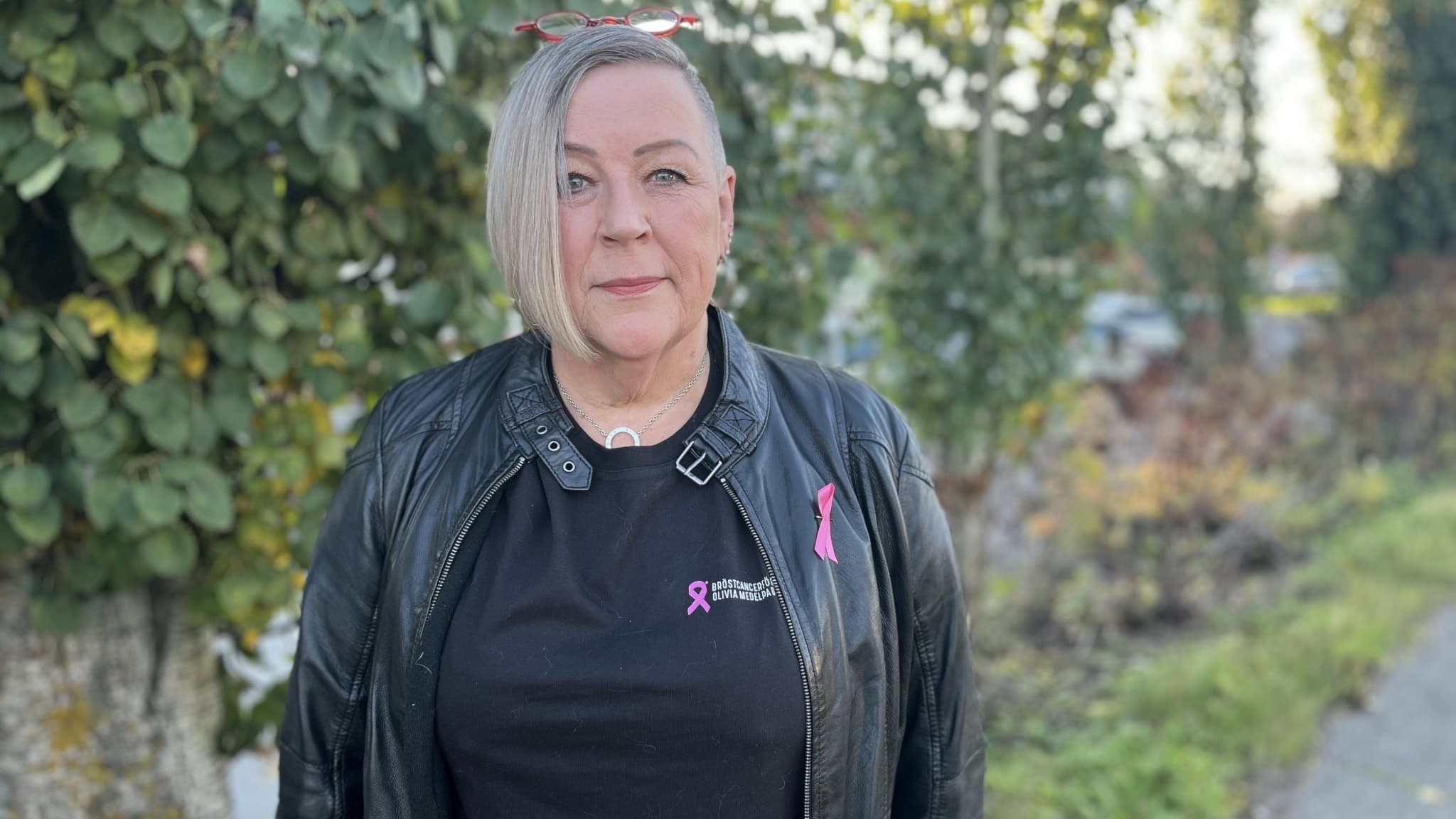 Tove Pallin, ordförande i bröstcancerföreningen Olivia i Medelpad, står utomhus framför grönskande buskar, klädd i en svart jacka och en tröja med rosa bröstcancersymboler.