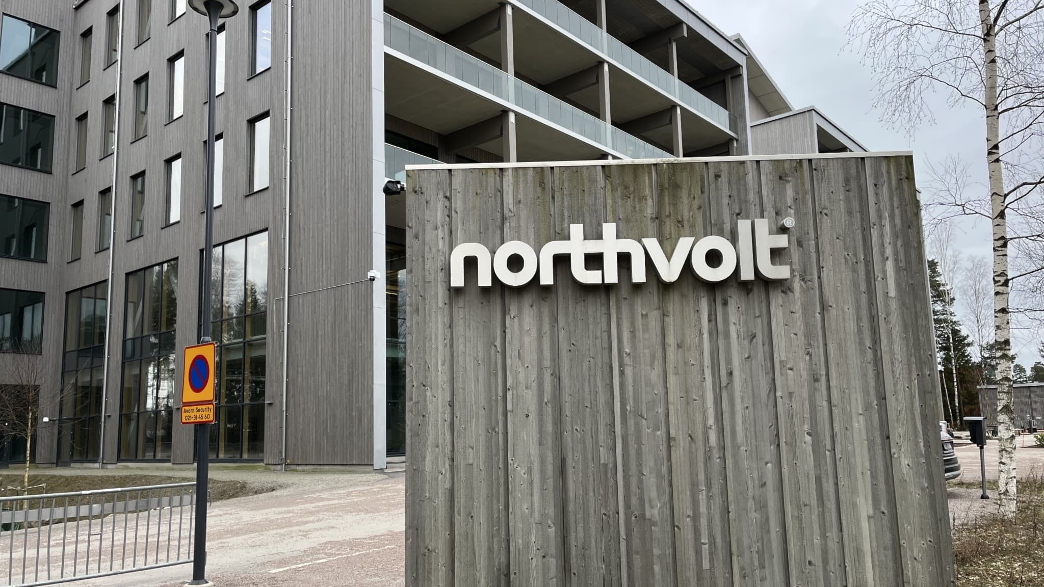 Bild av Northvolts byggnad i Västerås, med företagets logotyp på en betongvägg i förgrunden. Bakom syns moderna fasader och träd. En parkeringsskylt är placerad vid sidan av vägen.