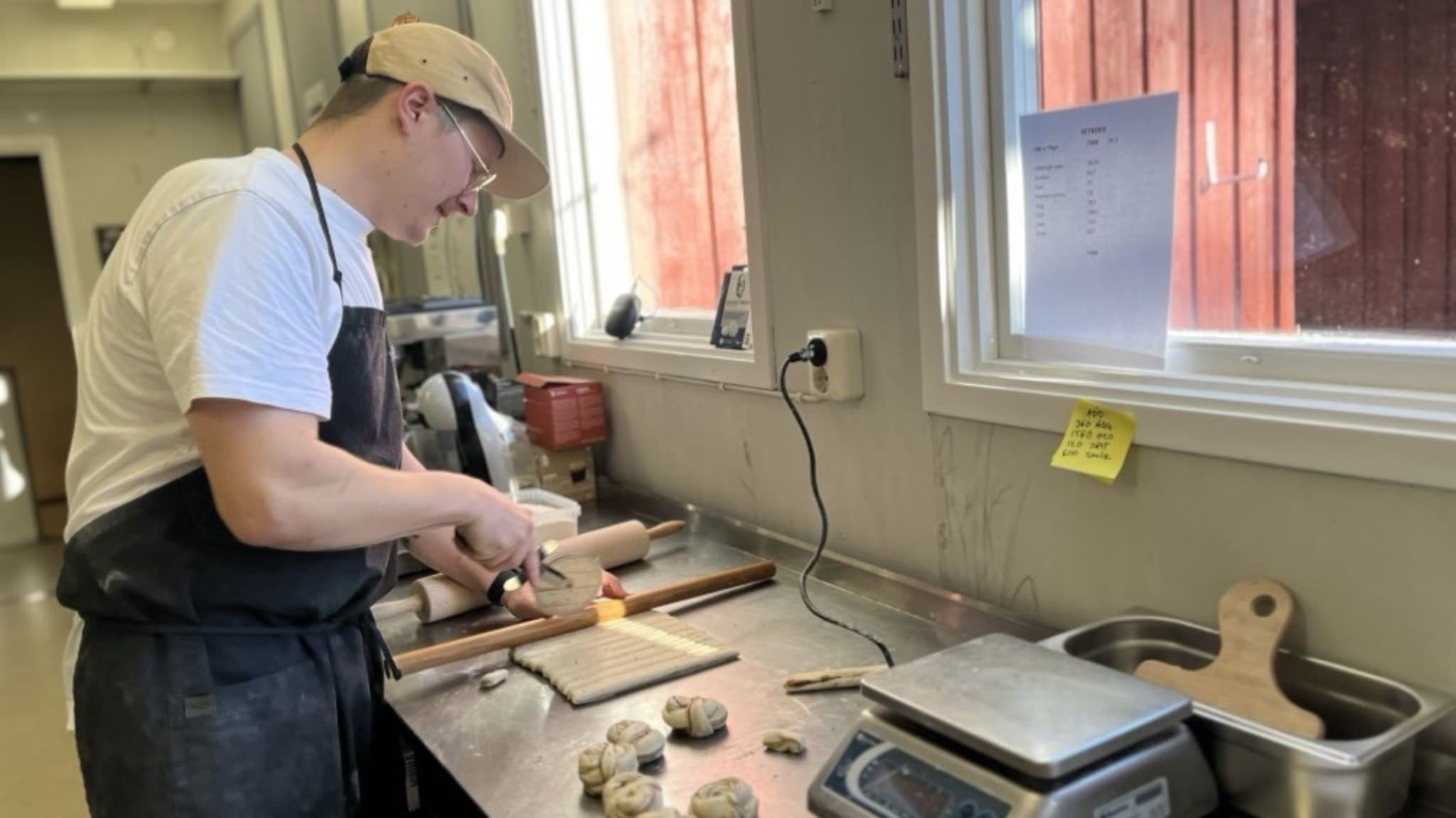 Bagaren Viktor Hed är i full gång med att baka kanelbullar. På bilden syns en man med förkläde, vit tröja och beige keps. Han står vid ett bakbord och gör kanelbullar. 