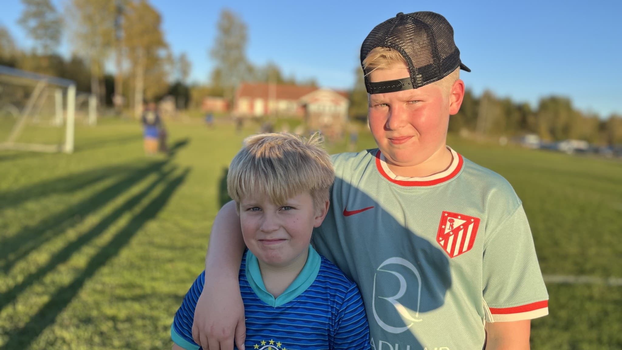 Bröderna Hugo och Walter Alriksson står på en fotbollsplan i kvällssol, klädda i fotbollströjor. Gräsplanen sträcker sig bakom dem med målburar och andra barn i bakgrunden.