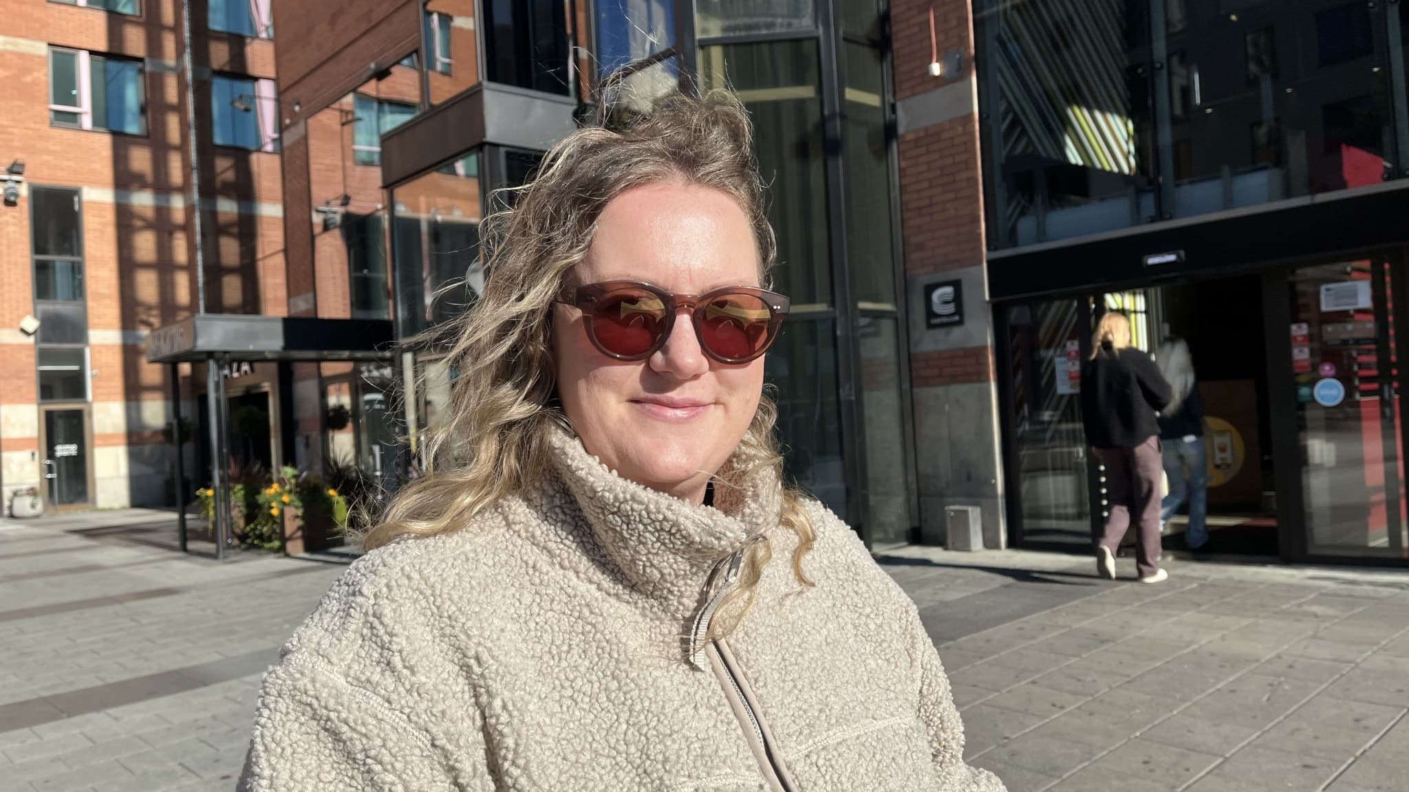 Alexandra Bjur står utomhus i centrala Västerås framför moderna byggnader i glas och tegel. Hon bär en ljus fleecejacka och det är soligt väder.
