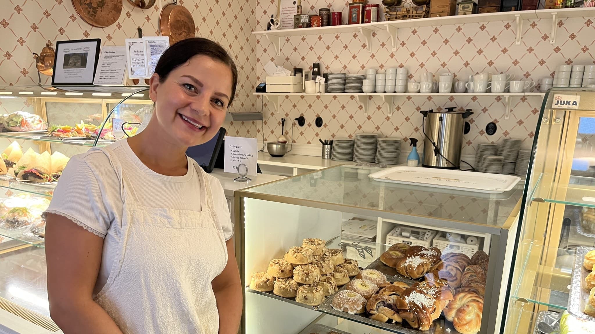 Jessica Bornström, delägare i Elsa Anderssons konditori i Norberg, står vid en glasmonter fylld med bakverk i ett klassiskt inrett café med ljusa väggar och hyllor med porslin.