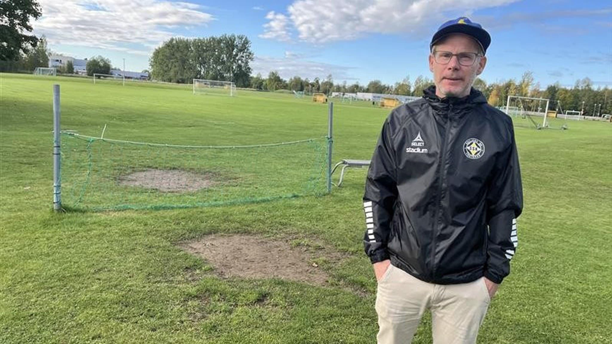 Lars ”Lalla” Kjellstrand från Skiljebo SK står på en gräsplan med fotbollsmål i bakgrunden, klädd i svart klubbjacka och keps. Soligt väder med blå himmel och moln.