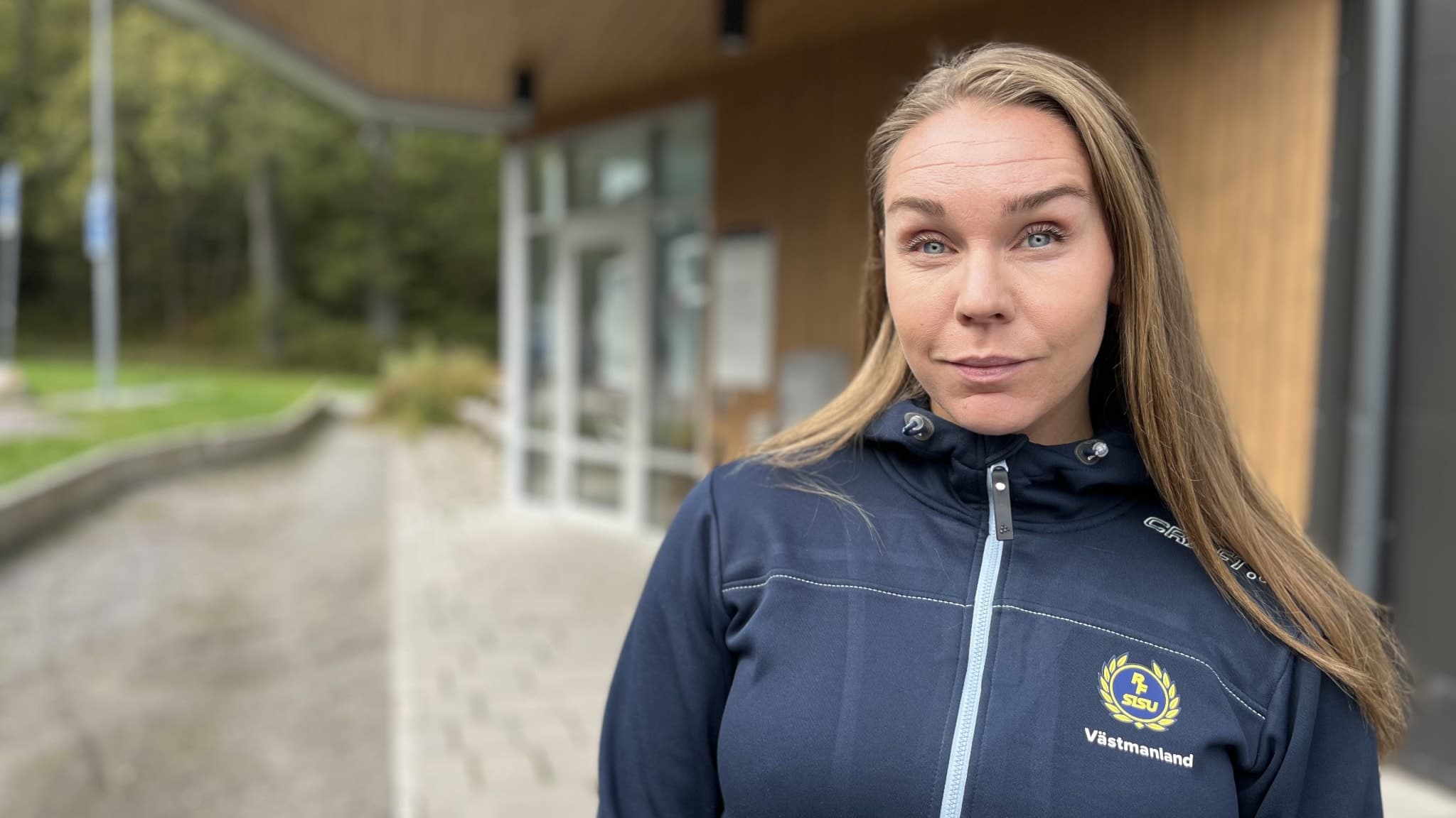 Person i mörkblå jacka med en gul logga står utanför en byggnad med glasfasad och träd i bakgrunden.
