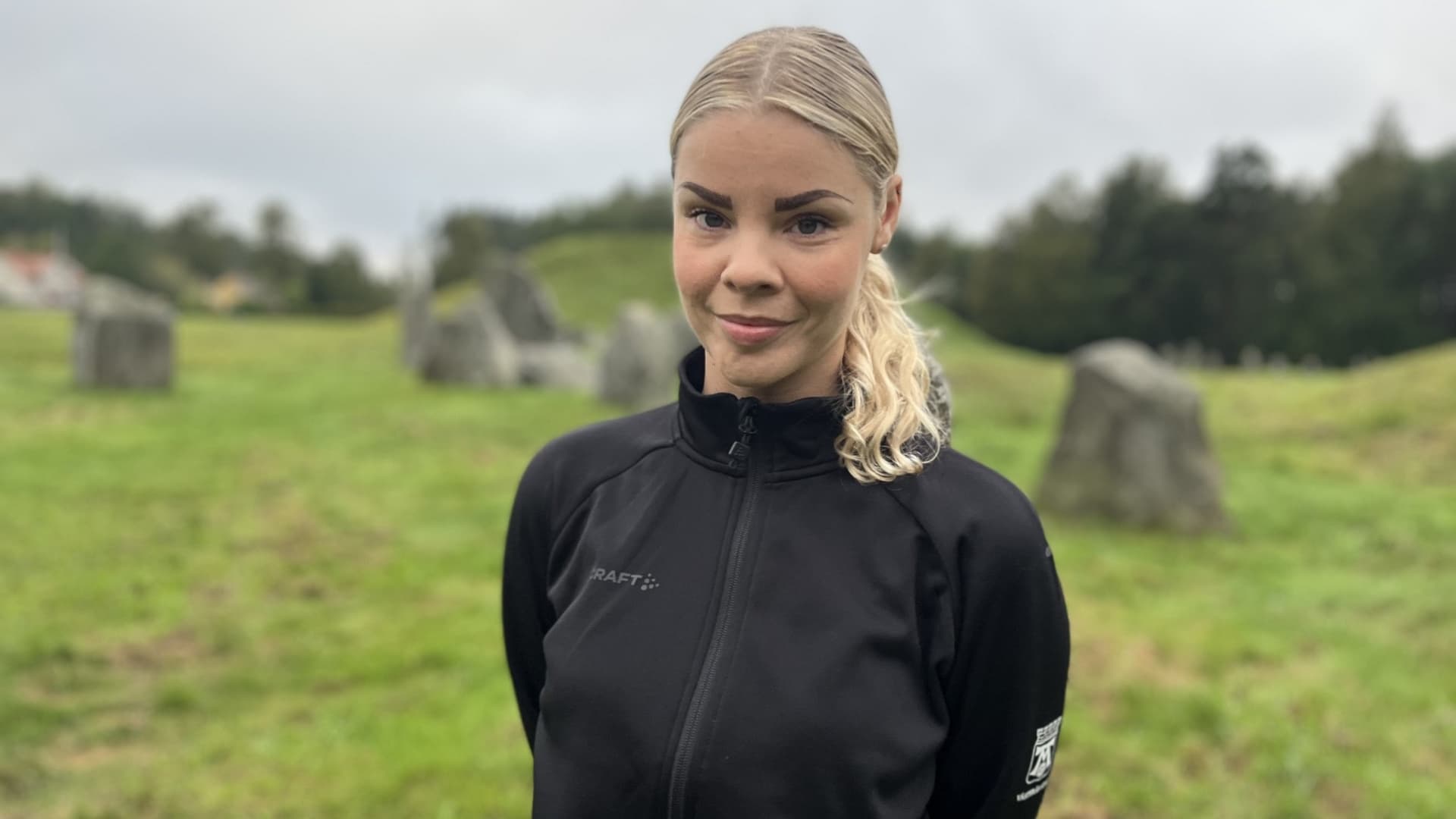 En person i svart jacka står på en grön äng omgiven av stenar, med träd i bakgrunden under en molnig himmel.