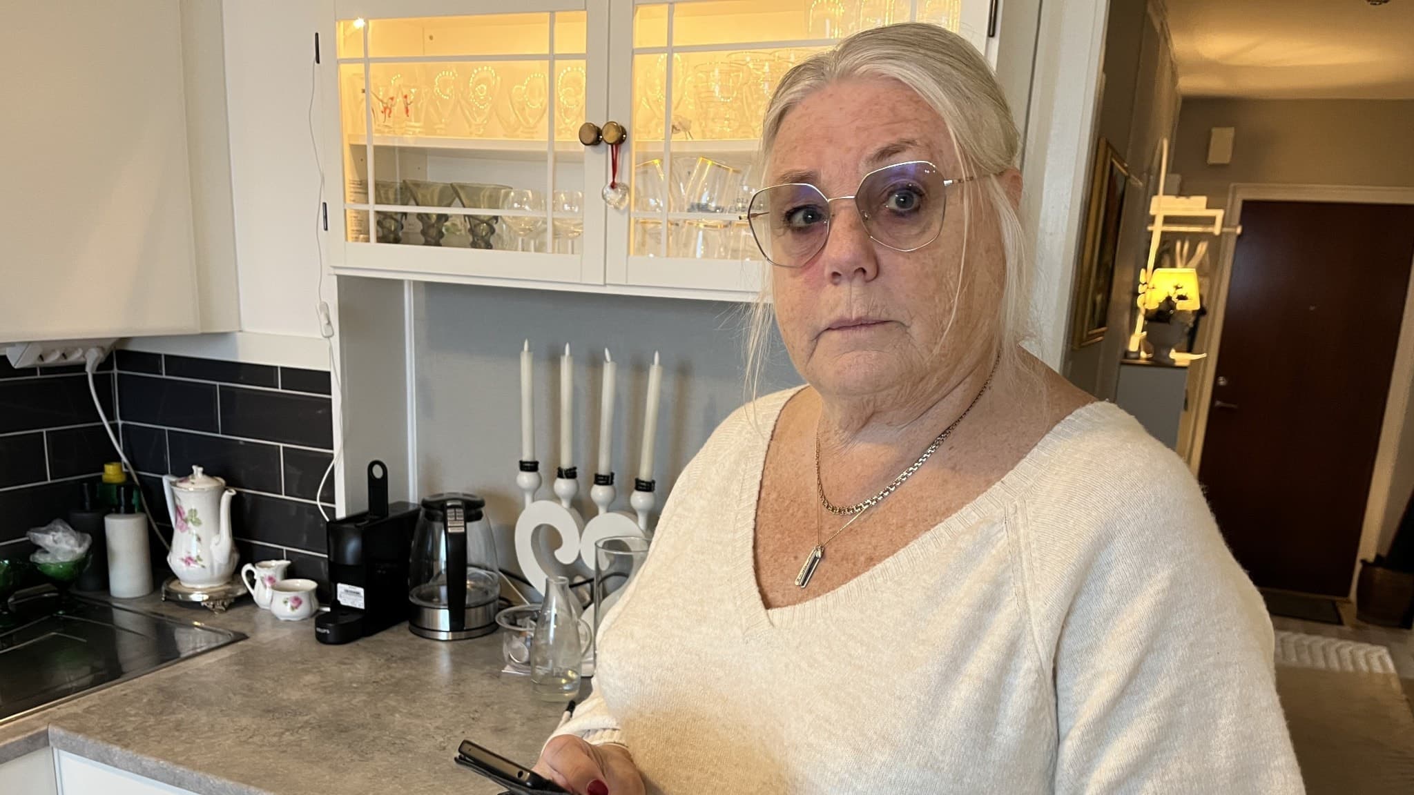 Eva-Lena Trondset, ordförande i PRO Fagersta och vice ordförande i PRO Västmanland, står i ett kök med vita skåp och svarta kakelplattor, hållandes en mobiltelefon.