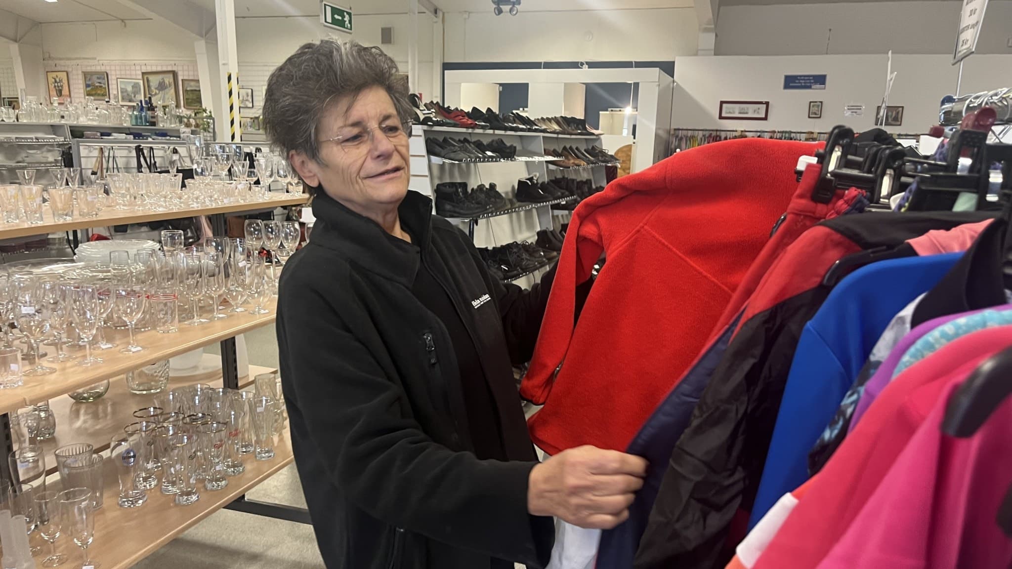 Mirjana Eriksson, lageransvarig på second hand-butiken Hela Människan i Sala, står bland klädhyllor och glasvaror och håller i en röd jacka, i en butiksmiljö.