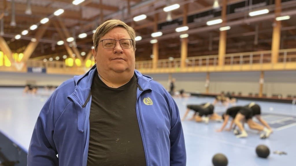 En person i blå jacka står framför en inomhushall där flera individer tränar med medicinbollar på en blå matta under tak med stark belysning.