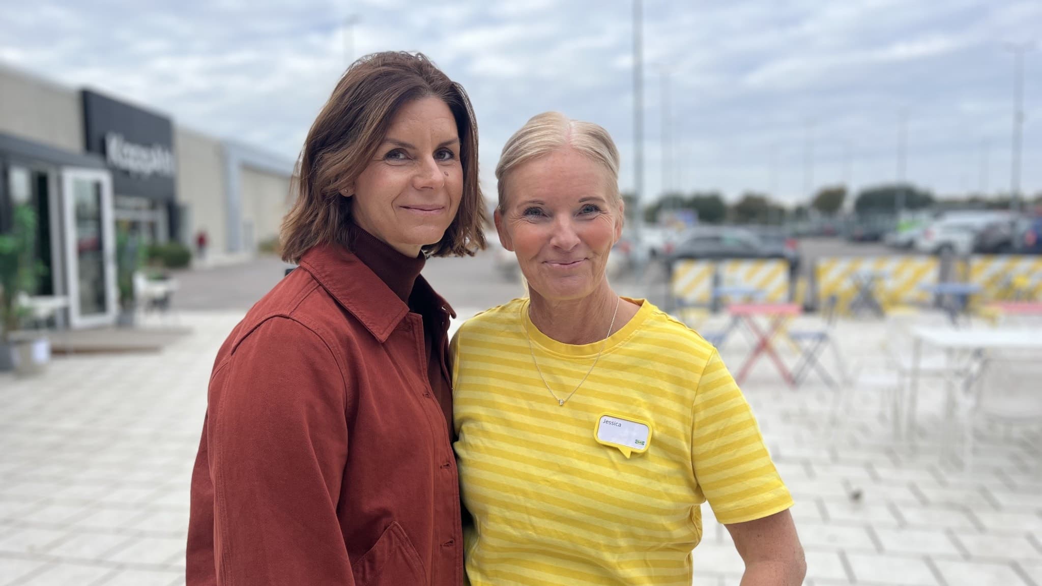 Två personer står utomhus på en stenlagd yta framför en byggnad med skyltar. Den ena har en röd skjorta, den andra en gul tröja. I bakgrunden syns bord, stolar och parkeringsplatser.