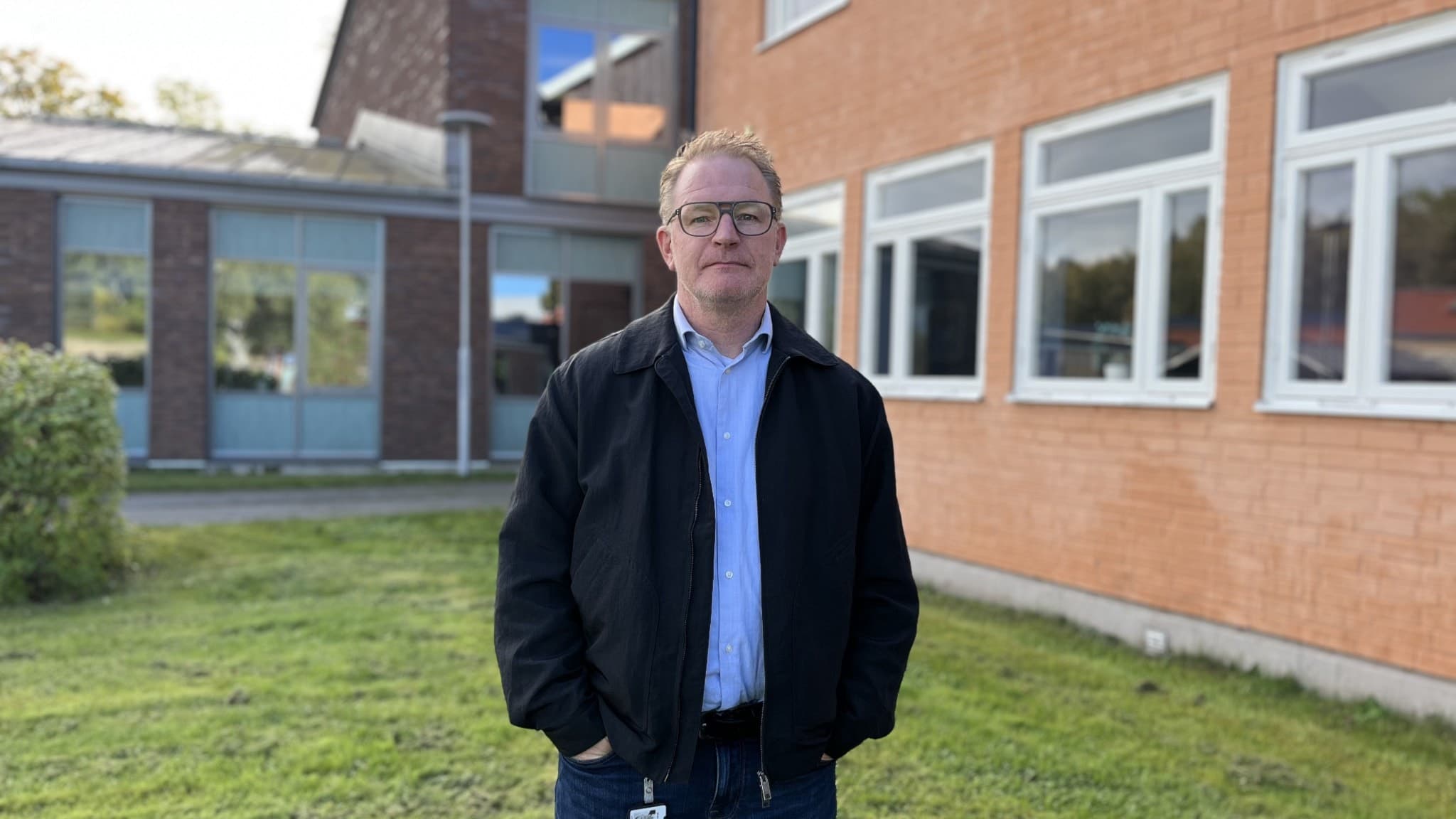 En person i mörk jacka och ljus skjorta står utomhus framför en tegelbyggnad med stora fönster. Grön gräsmatta omger byggnaden under en klar himmel.
