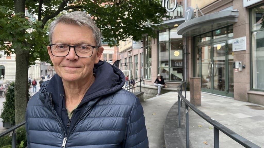 Mats Lindblad står utomhus framför en byggnad med glasfasad och skyltar. Han bär en mörkblå jacka och det är träd i bakgrunden. Miljön ser ut som en stadsmiljö med trottoar och räcken.