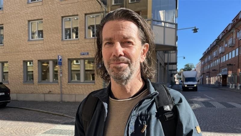 Lars Wallin står utomhus framför en tegelbyggnad i Uddevalla. Han bär en mörk jacka och ryggsäck. Solen lyser på gatan med parkerade bilar och gångtrafik i bakgrunden.