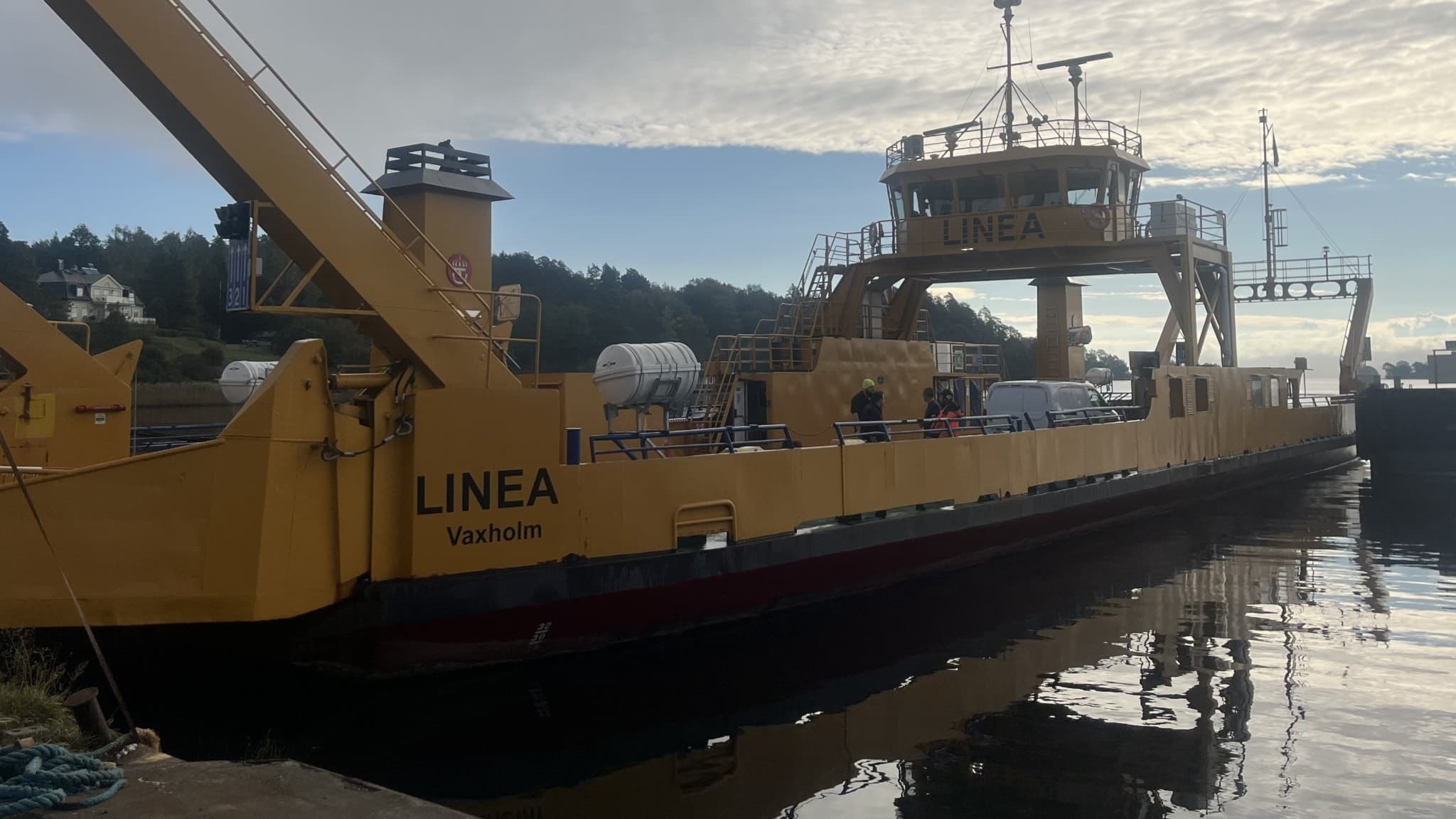 Gul färja vid kajen, namngiven Linea, som ska ersätta Dalbobron under dess stängning. Färjan ligger stilla i vatten med skog och molnig himmel i bakgrunden.
