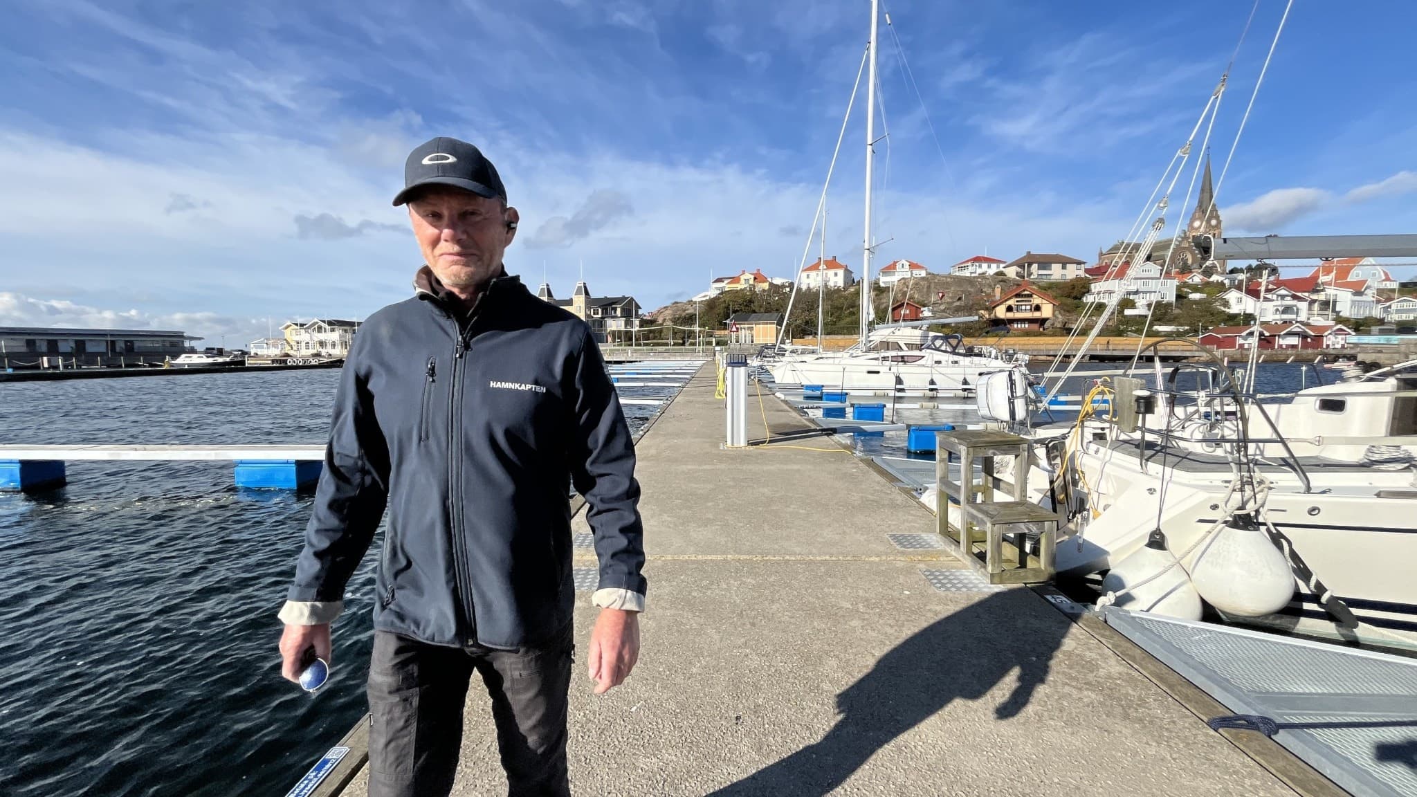 Kent Håkonsen, hamnkapten i Lysekil, står vid en brygga med båtar förtöjda och en vy över staden i bakgrunden. Solen skiner och himlen är blå med lätta moln.