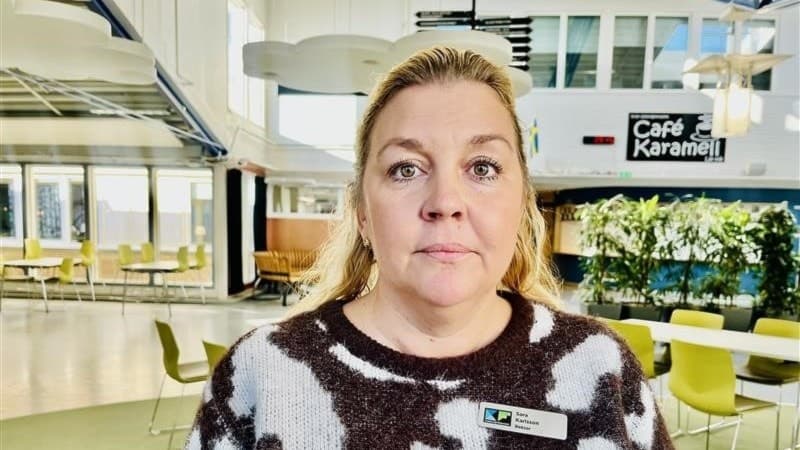 En person med blont hår som är rektor på skolan, står inomhus i en ljus och modern miljö med cafébord och växter i bakgrunden. Skylten 'Café Karamell' syns på väggen.