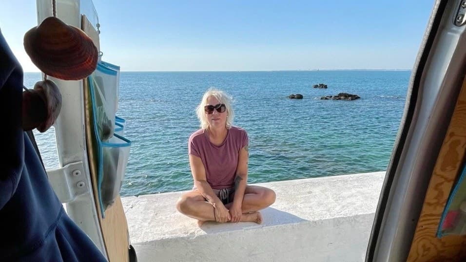 Bettina Axelsson sitter avslappnat i solen på en vit stenmur vid havet, med blå himmel och klart vatten i bakgrunden, fotograferad från insidan av en bil.