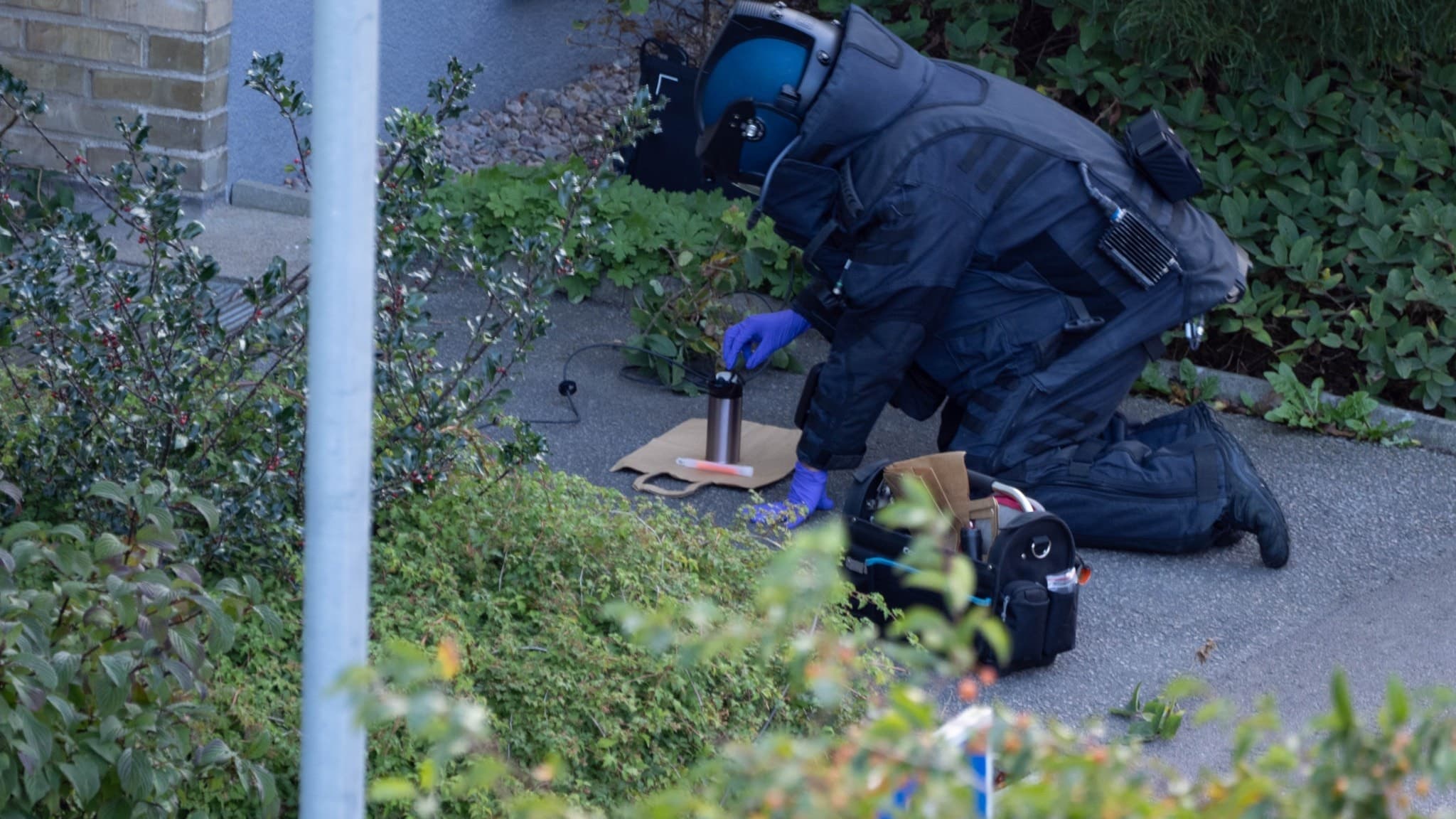 En bombtekniker i skyddsutrustning undersöker en misstänkt termosbomb utomhus, omgiven av grönska och byggnadsmaterial.