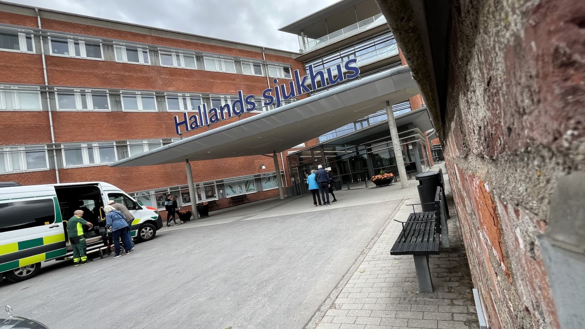 Entrén till Hallands sjukhus Halmstad med en ambulans parkerad utanför. Människor rör sig vid ingången under en molnig himmel. Tegelbyggnad och bänkar syns i förgrunden.