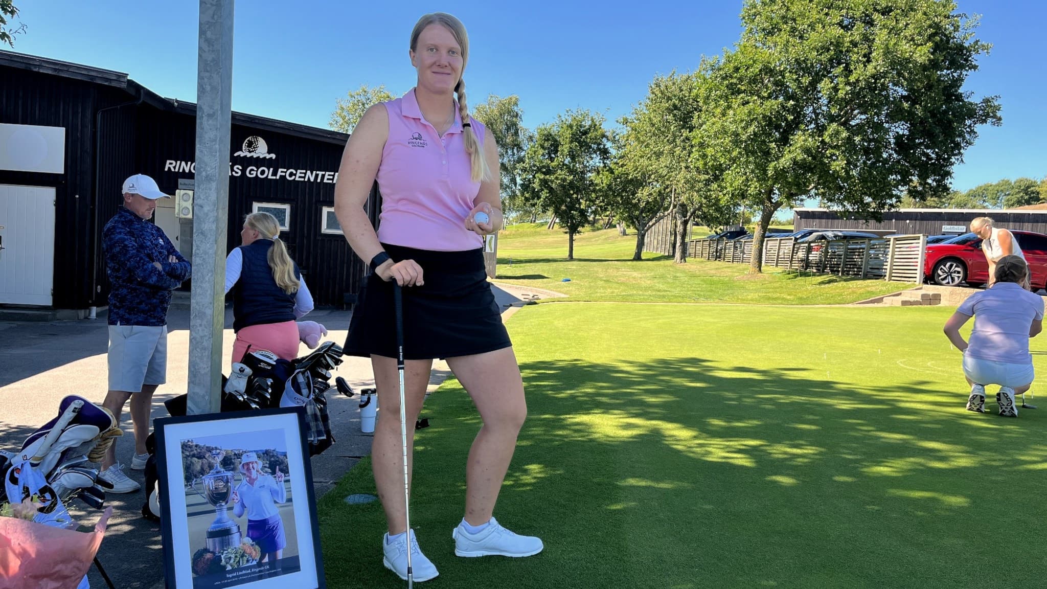 Ingrid Lindblad står med en golfklubba på en grön golfbana, klädd i en lila topp och svart kjol. I bakgrunden syns andra golfspelare och en tavla med en golfrelaterad bild.