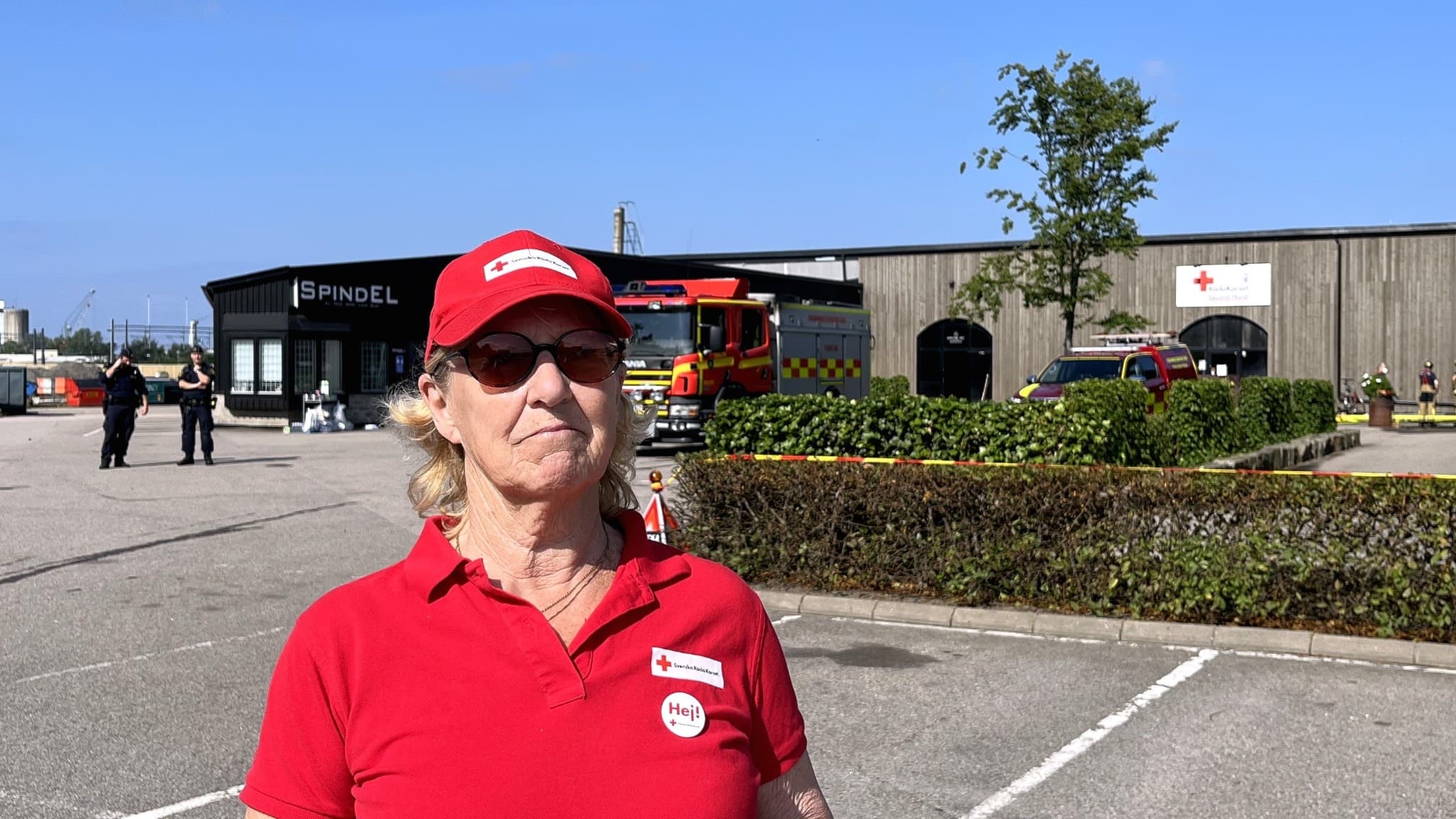 En kvinna i röd pikétröja och keps står framför en butikslokal som är avspärrad efter en brand. Brandbilar och räddningspersonal syns vid den totalförstörda byggnaden.