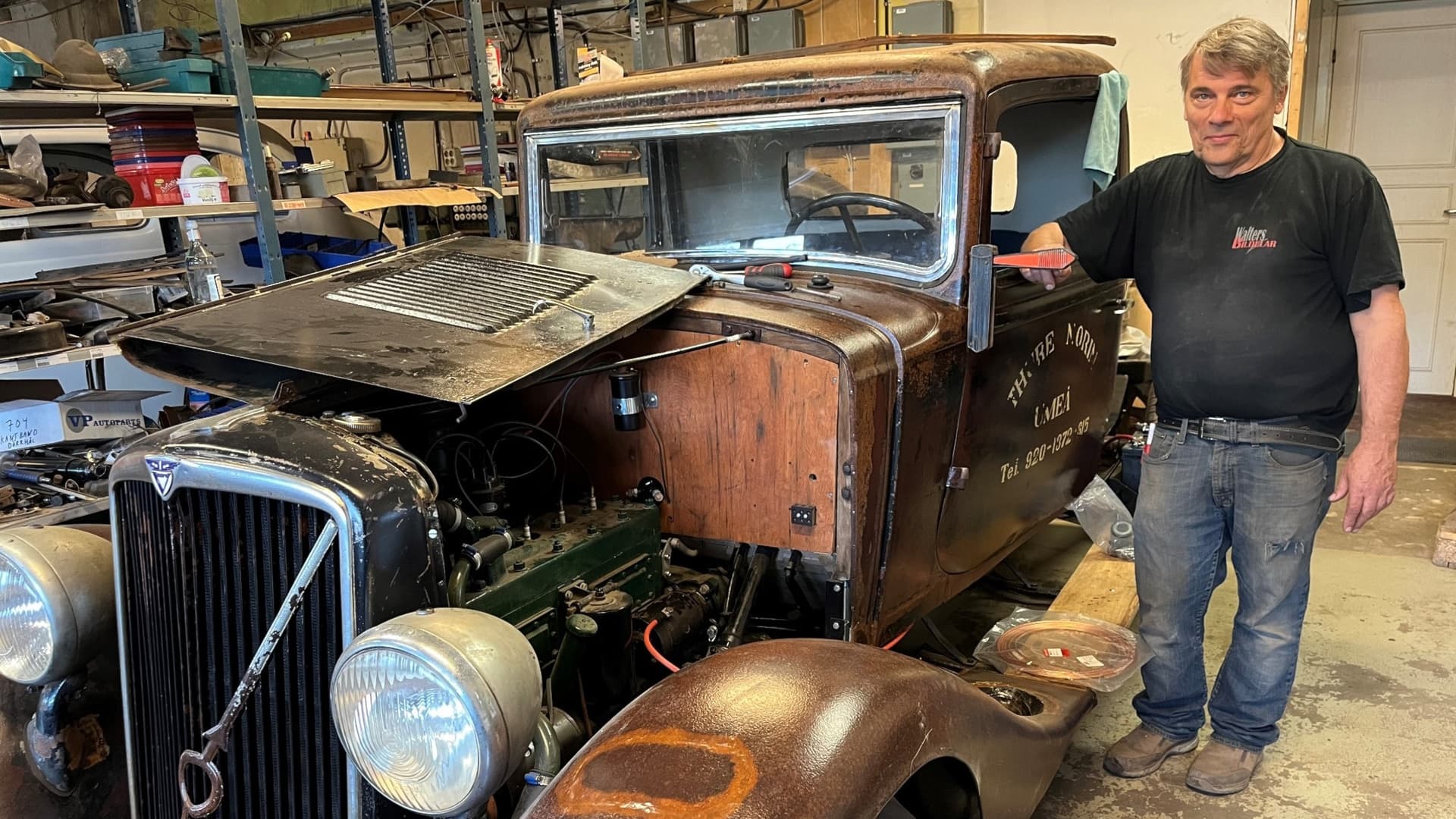 En person står bredvid en klassisk bil under restaurering i en verkstad. Bilen har rostiga detaljer och en öppen motorhuv som avslöjar arbetet på motorn.