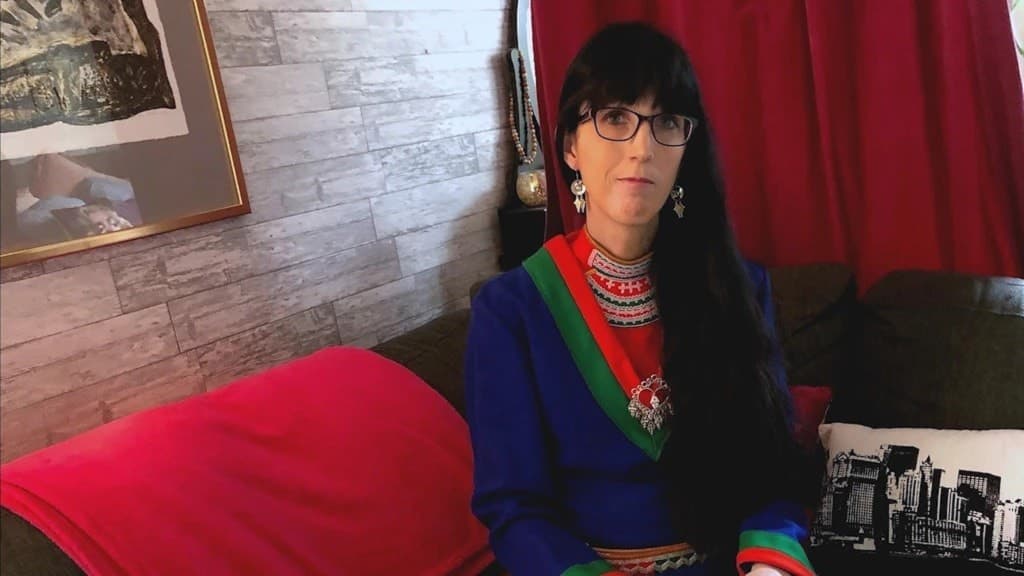 Eleonora Marsfjell sitter på en soffa klädd i traditionell samisk dräkt med färgglada detaljer. Bakgrunden består av en trävägg, en tavla och röda gardiner.