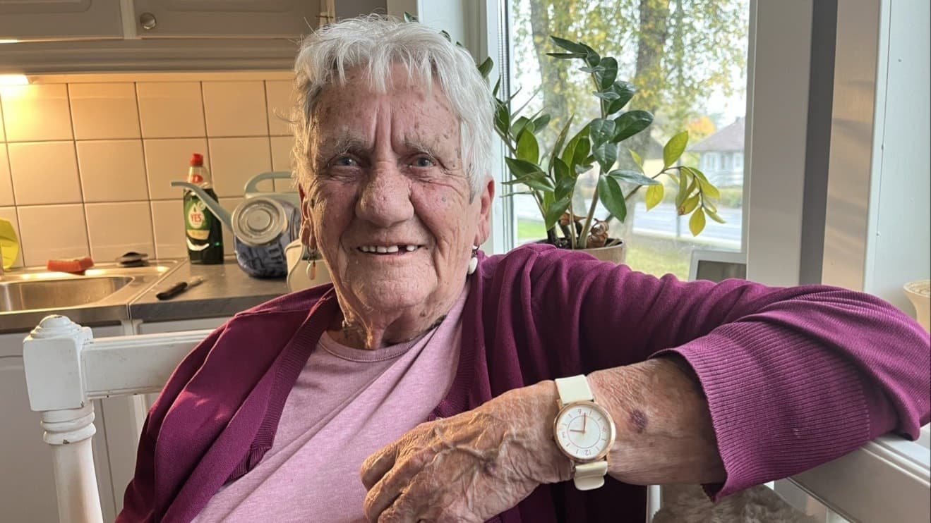 En äldre kvinna sitter i ett kök med en rosa tröja och vinröd kofta. Hennes hand med en vit klocka vilar på fönsterblecket.